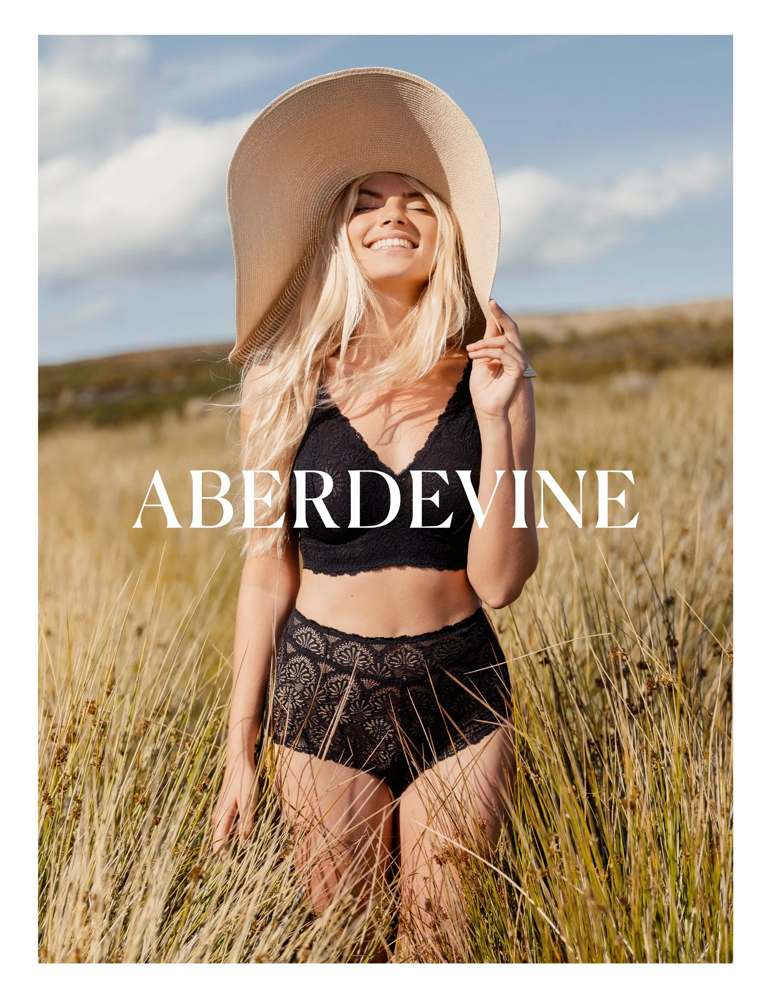 A woman smiling and wearing a wide-brimmed sunhat and black lace lingerie, standing in a field of tall grass with a partly cloudy sky.