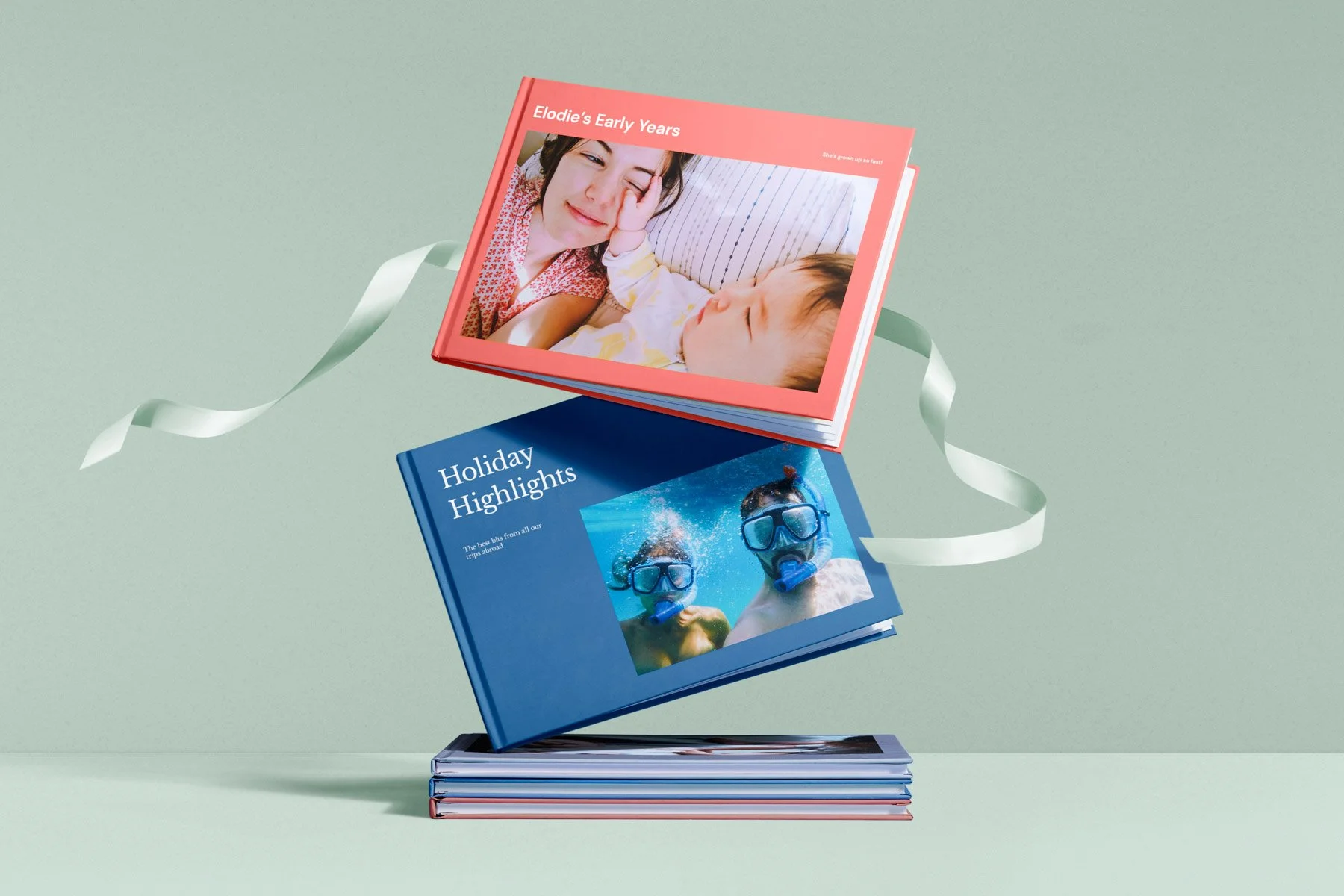 A stack of two colorful photo books with a third open book on top, displaying pictures of children and travel scenes against a mint green background.