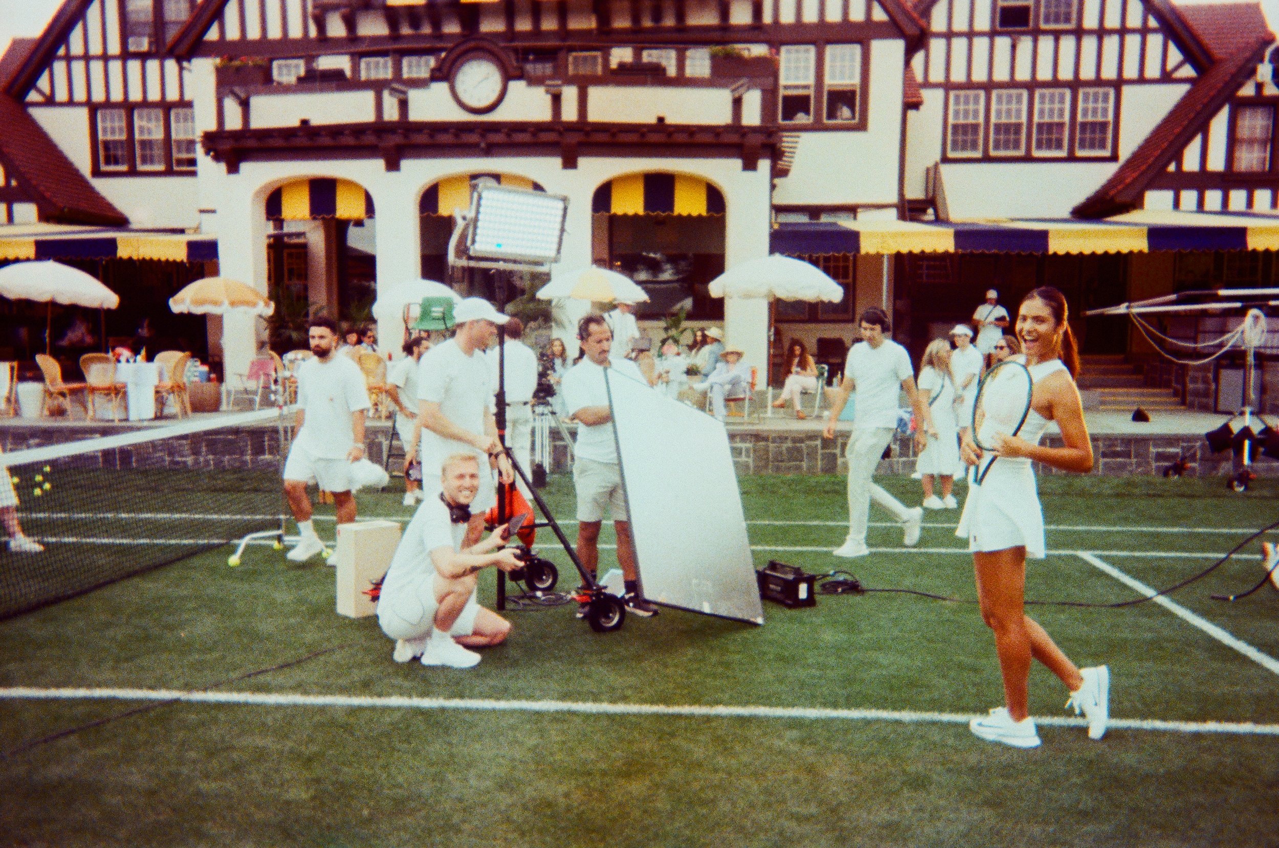 Group of people on a tennis court preparing for a shoot, some holding tennis rackets, with a building and spectators in the background.