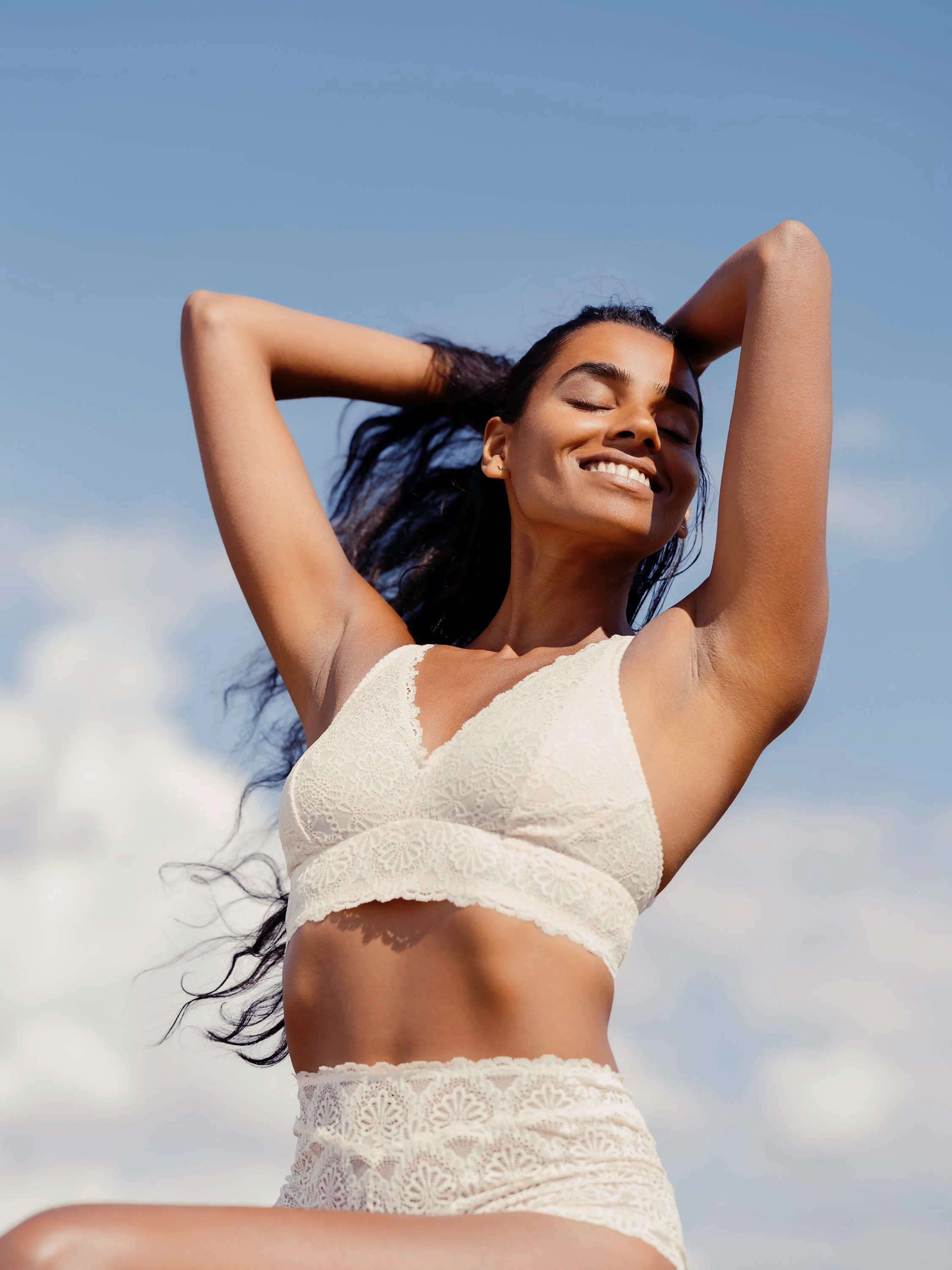 Smiling woman with dark hair in a lace white bralette and high-waisted lace shorts, standing outdoors with arms raised behind her head and eyes closed, against a partly cloudy blue sky.
