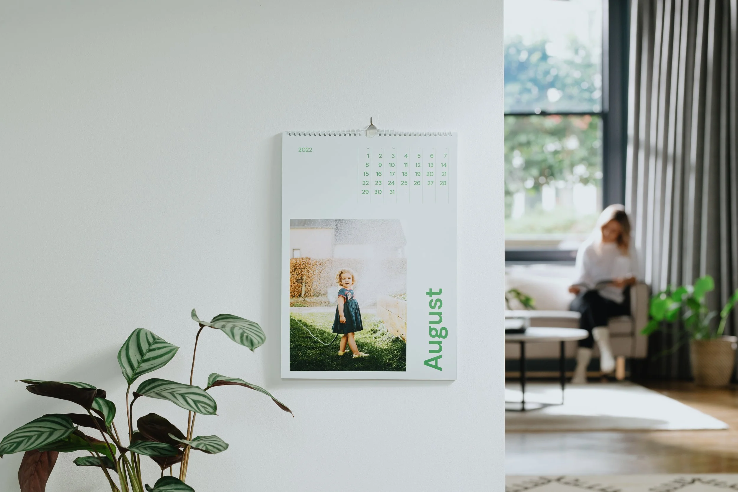 A calendar on a white wall showing August 2022, with a photo of a young girl in a navy dress watering plants outdoors. In the background, a woman is sitting on a sofa near a large window, reading a book in a bright, modern living room.