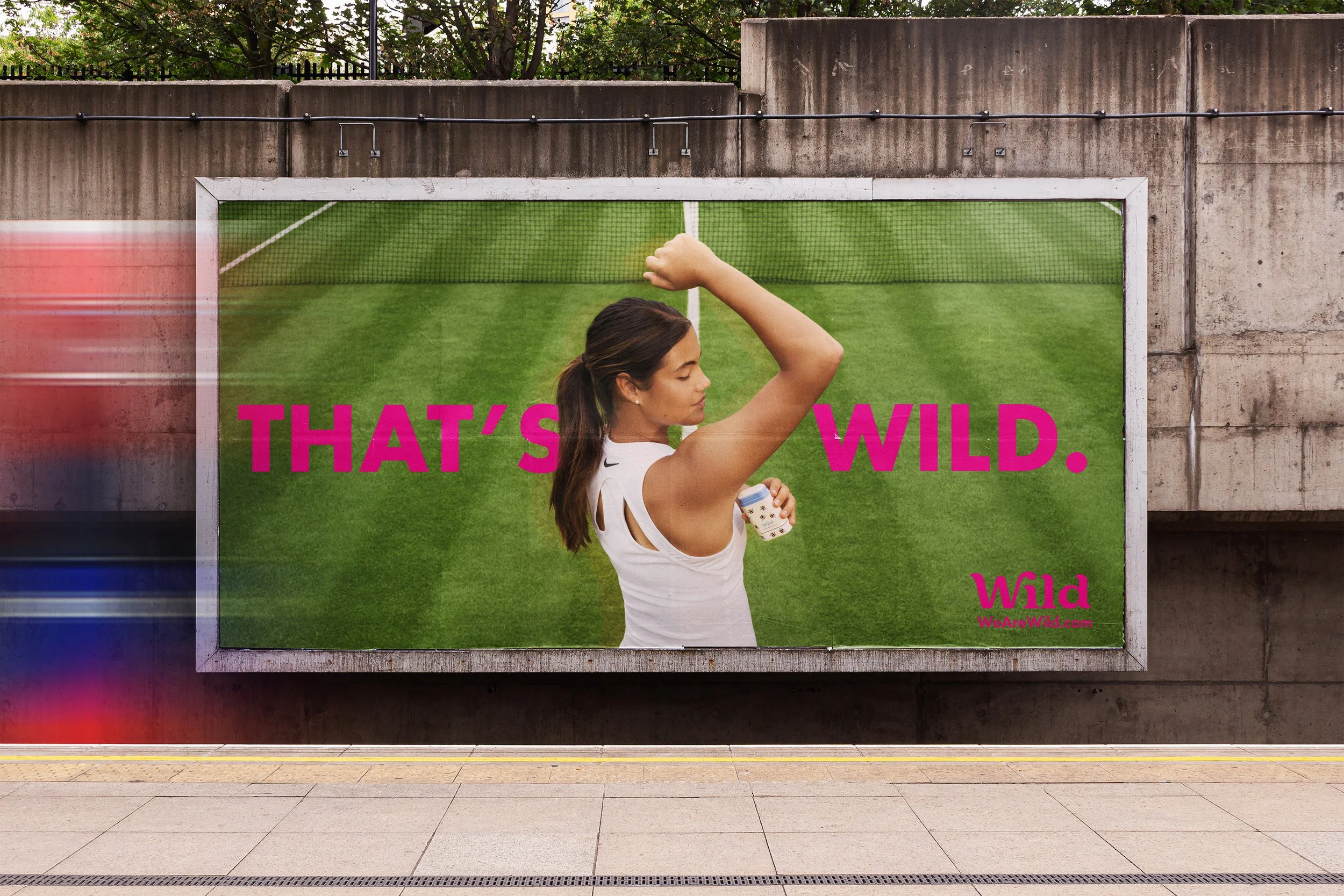 A woman with long dark hair in a ponytail wearing a white tank top holding a beverage cup, standing in front of a green grass background with the bold pink text 'THAT'S WILD.' and the smaller pink text 'Wild WeAreWild.com'.