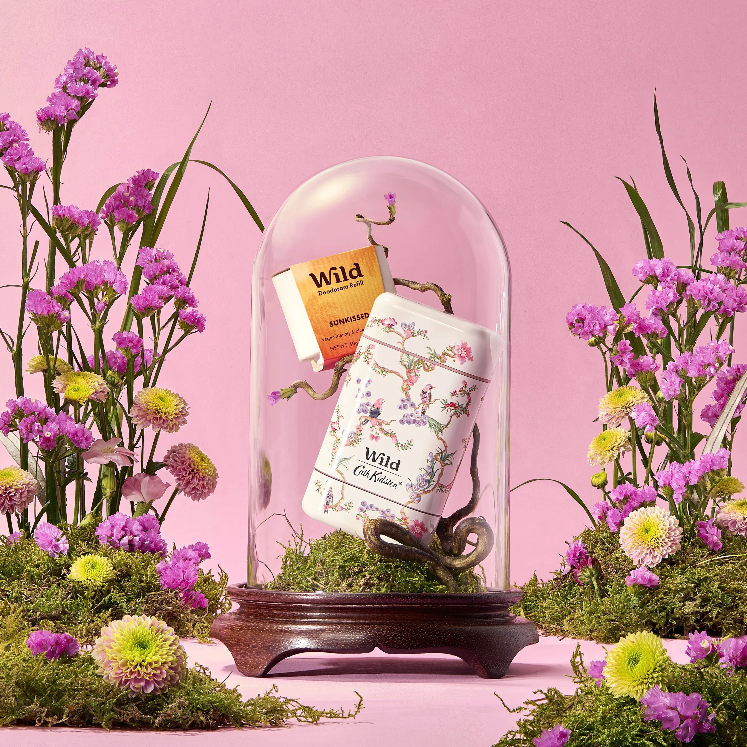 Decorative display of Wild brand toiletries including a floral patterned container and a deodorant refill bottle, surrounded by pink and yellow flowers on mossy ground with a pink background.