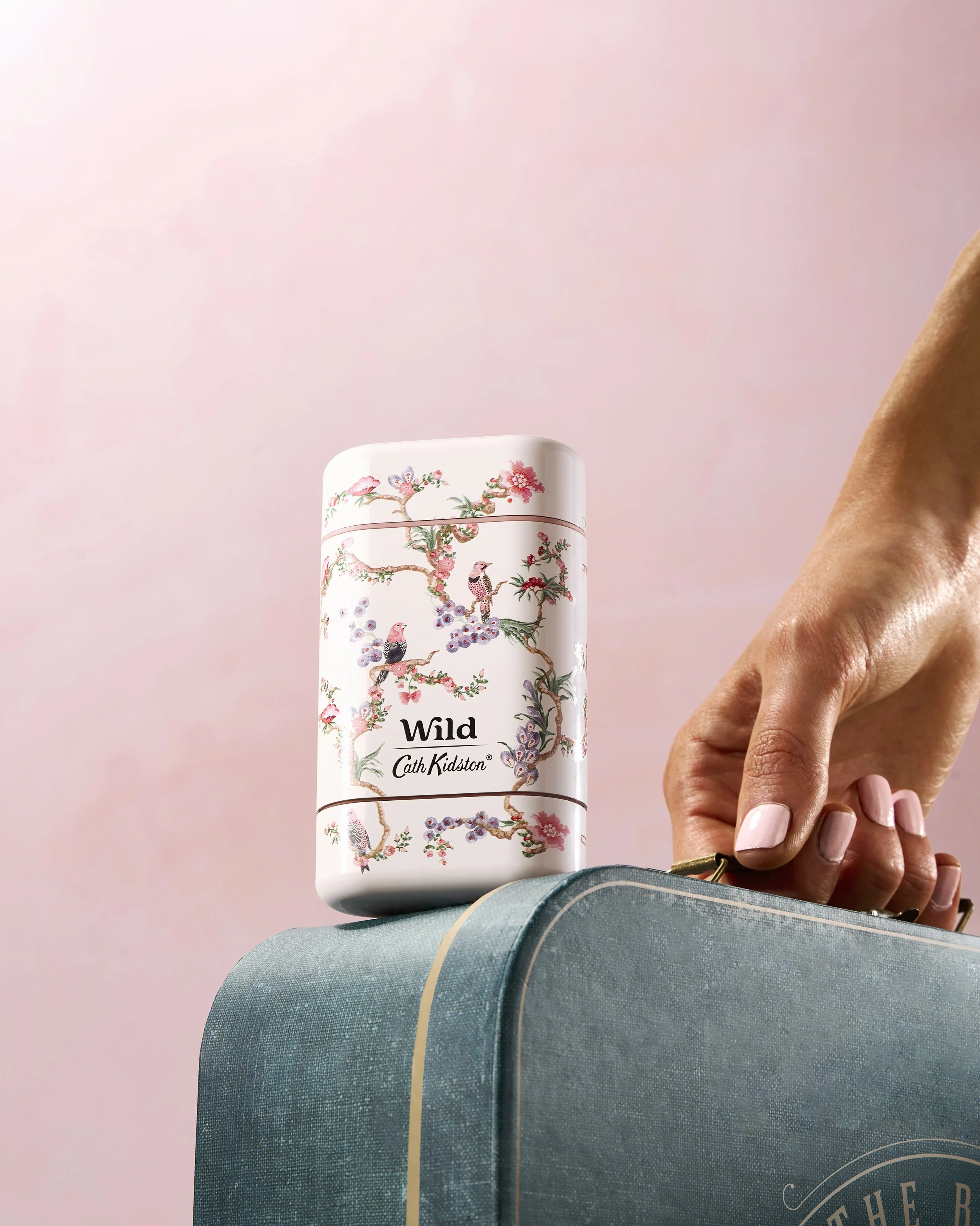 A hand reaching to open a teal suitcase with a white tins containing birds and flowers on a pink background.