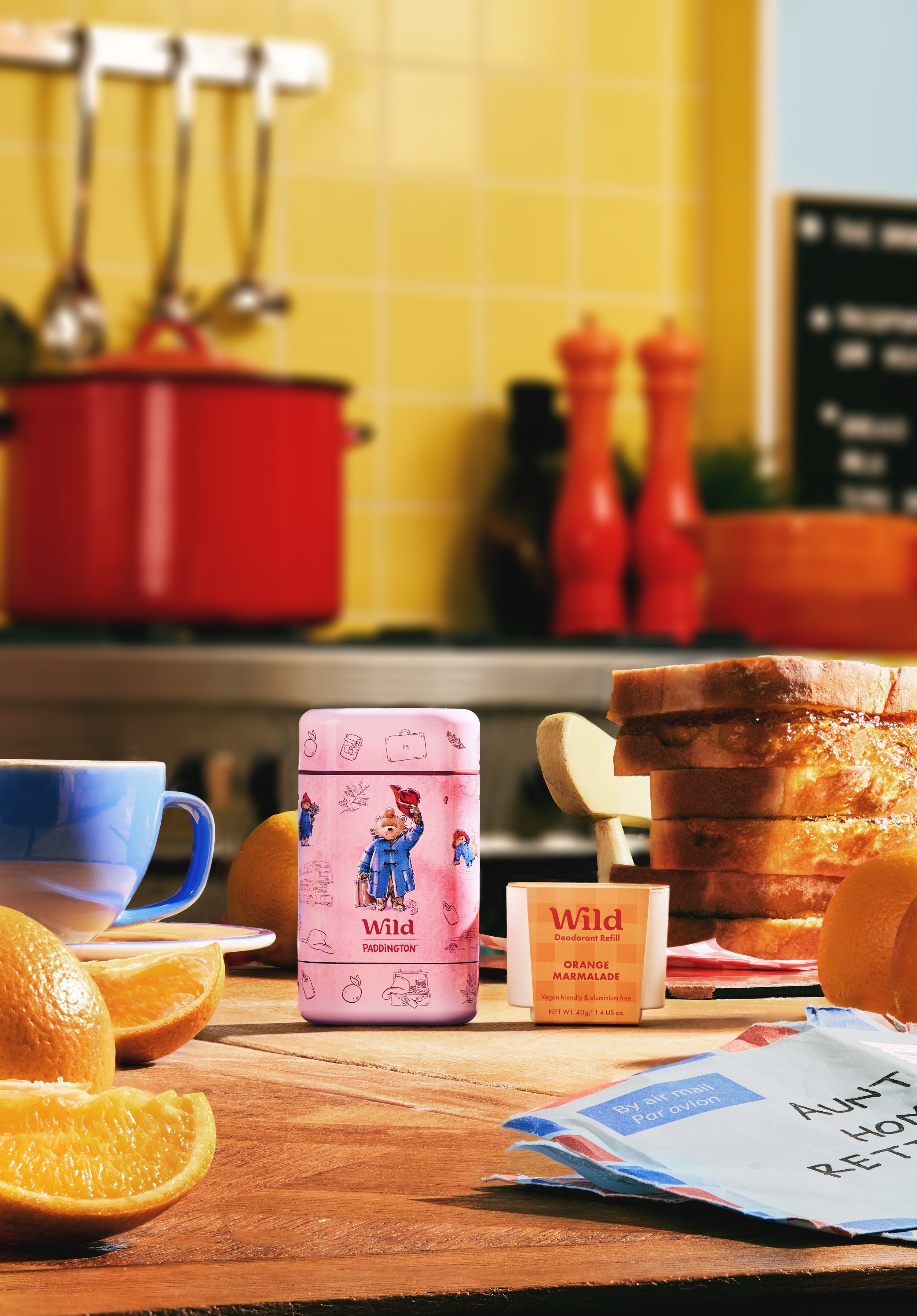A stack of sliced toasted bread, oranges, a blue mug filled with tea, a pink tin labeled 'Wild Paddington,' a small container of orange marmalade, a handwritten note, and various kitchen items in the background.