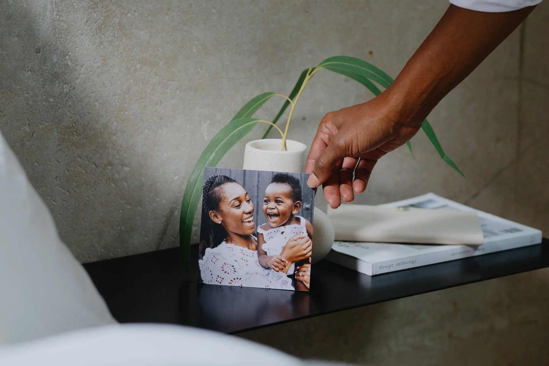 A person's hand holding a photo of a woman and a young girl smiling at each other, placed on a black surface near a potted plant with green leaves and a white ceramic vase, with books in the background.