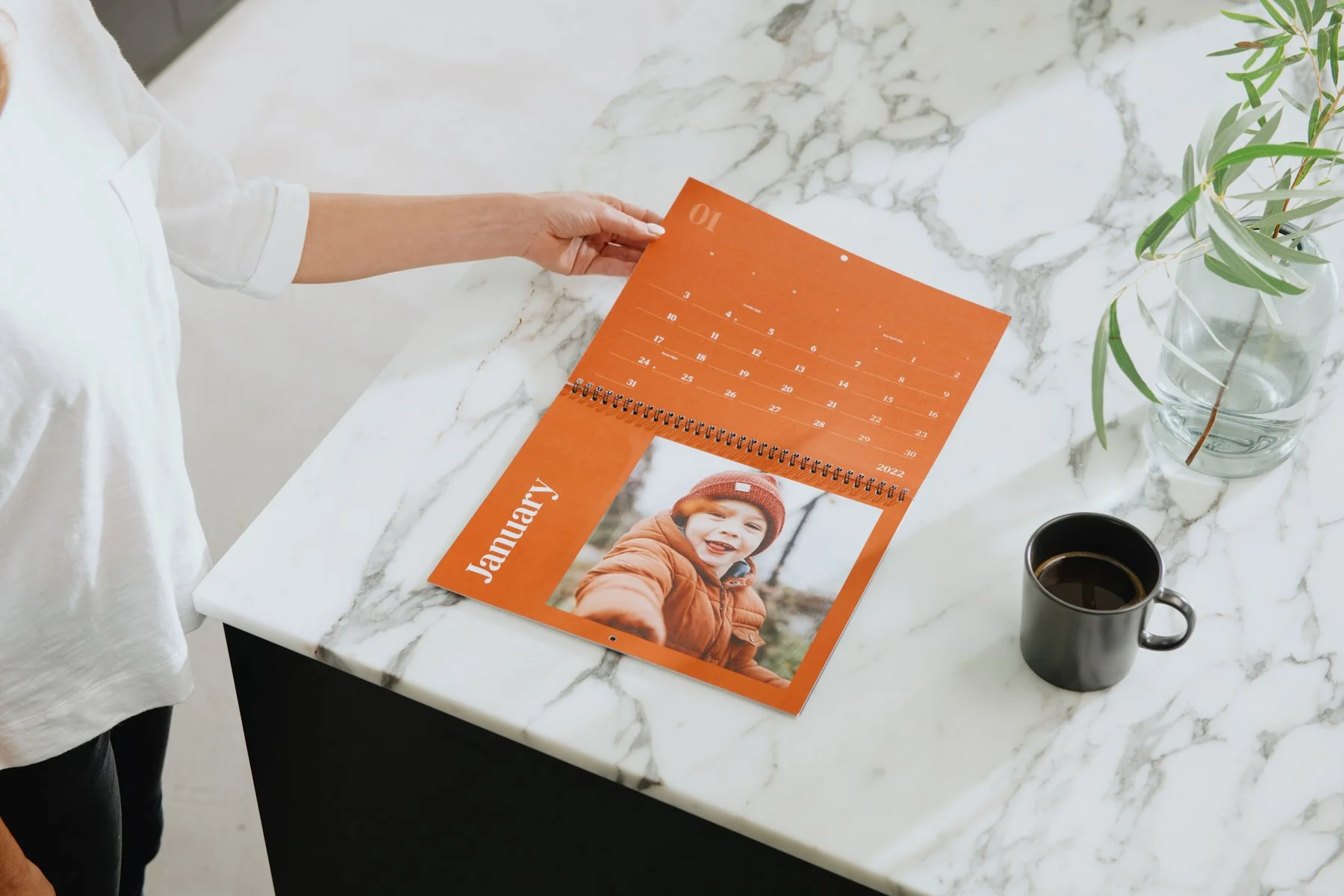 A person holding an orange January 2023 calendar with a photo of a smiling boy in a brown coat and red hat on a white marble table. Nearby are a black coffee mug and a glass vase with green leaves.