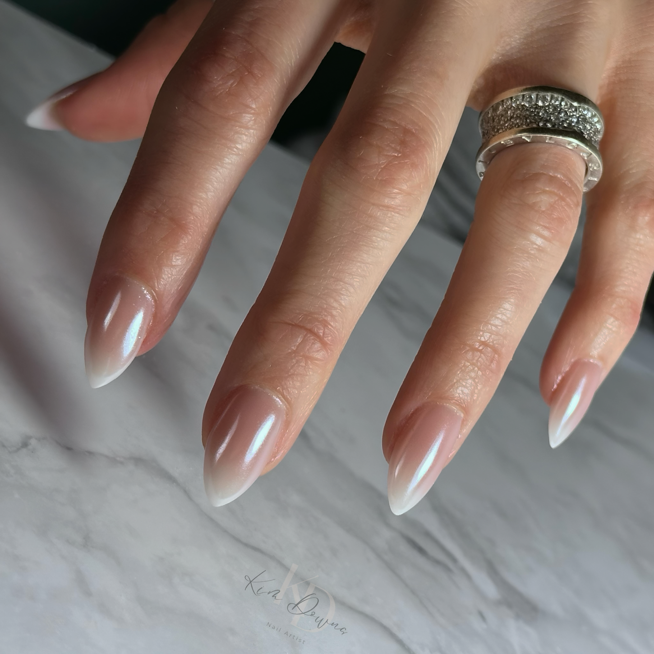 Close-up of a hand with long, glossy, French ombre nails and two silver rings with rhinestones on the ring finger, resting on a white textured surface.
