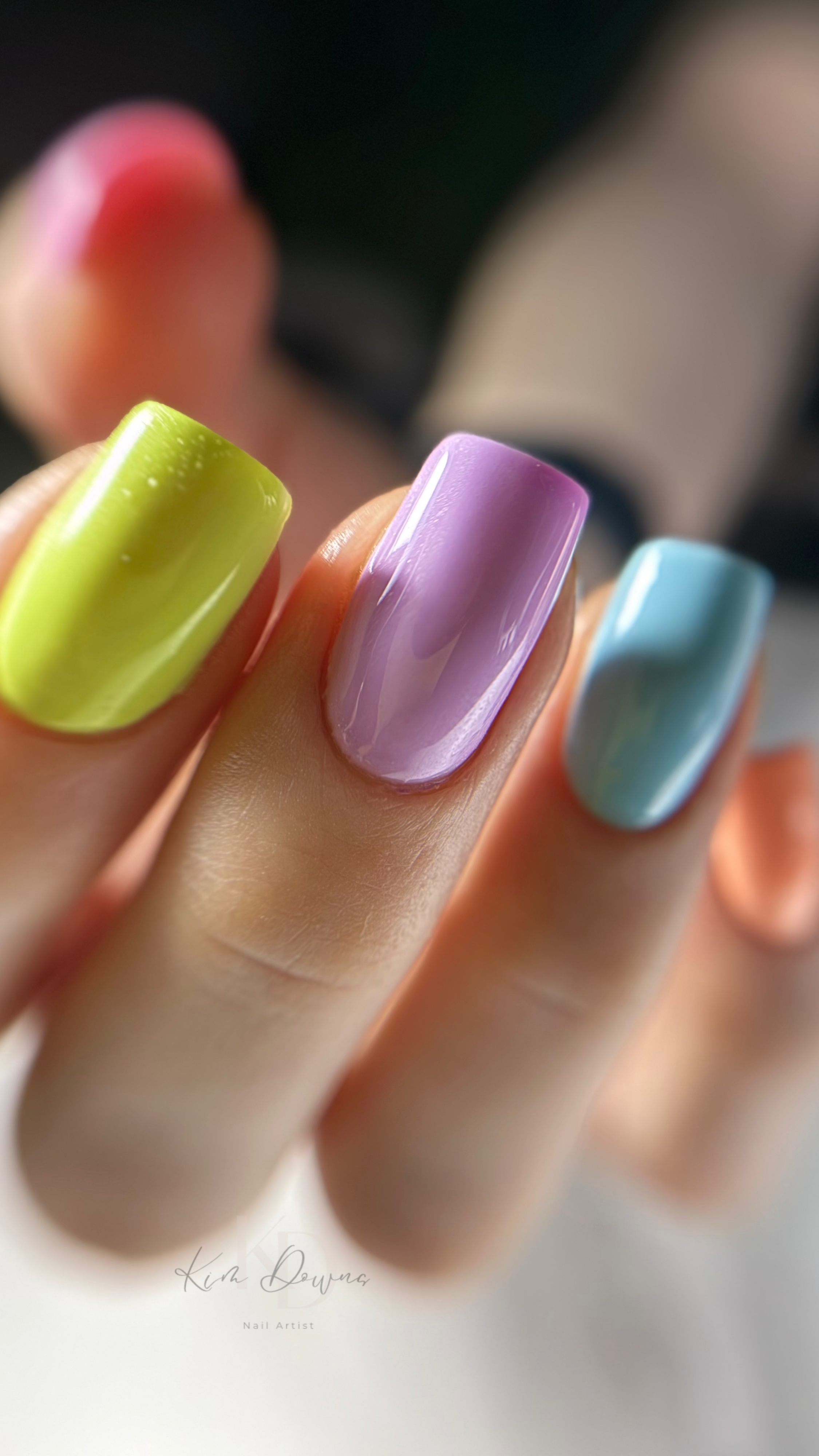Close-up of a hand with colorful pastel nail polish on each fingernail, including green, purple, and blue shades.