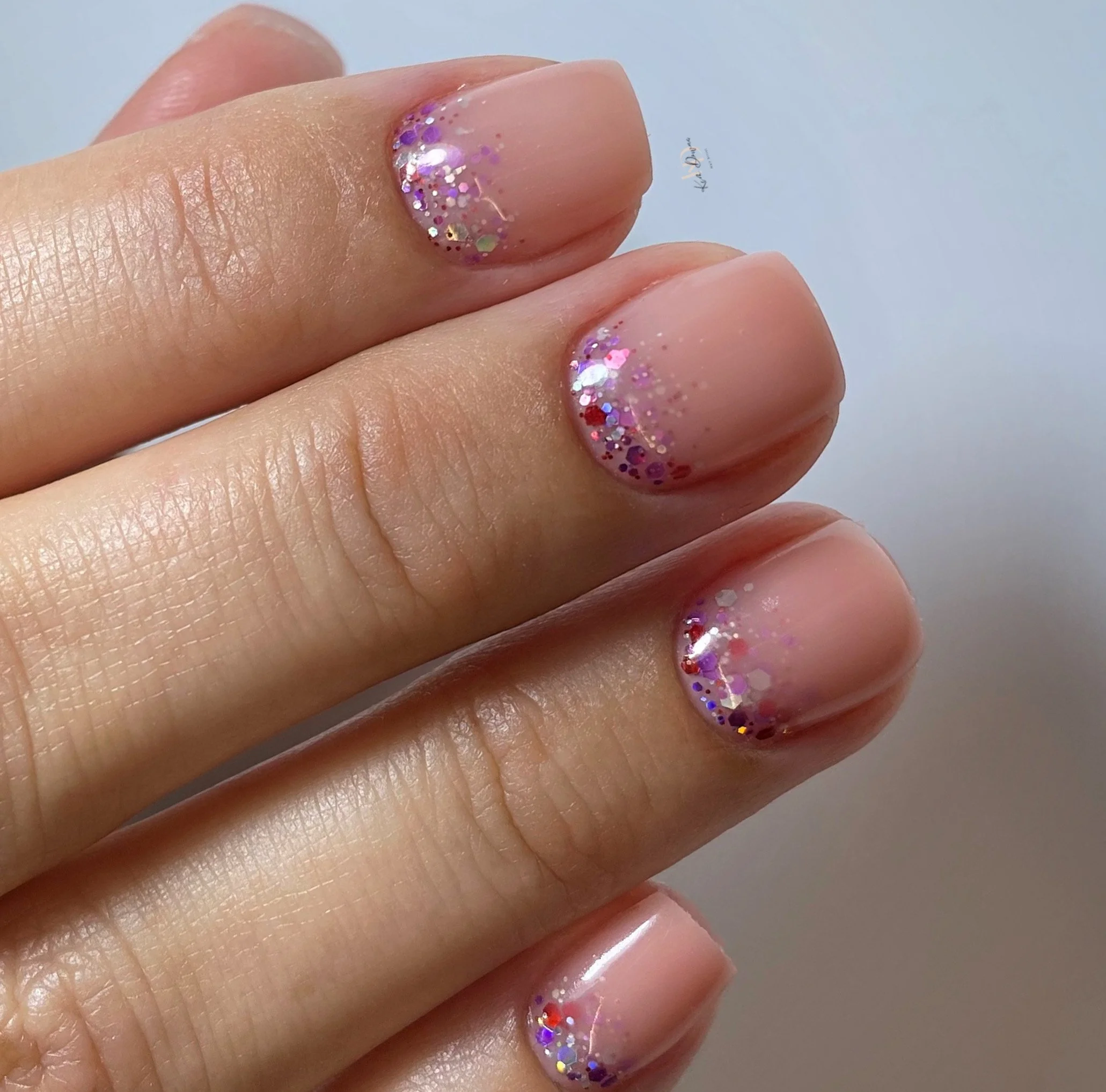 Close-up of a hand with manicured nails featuring nude polish with glitter accents at the tips.