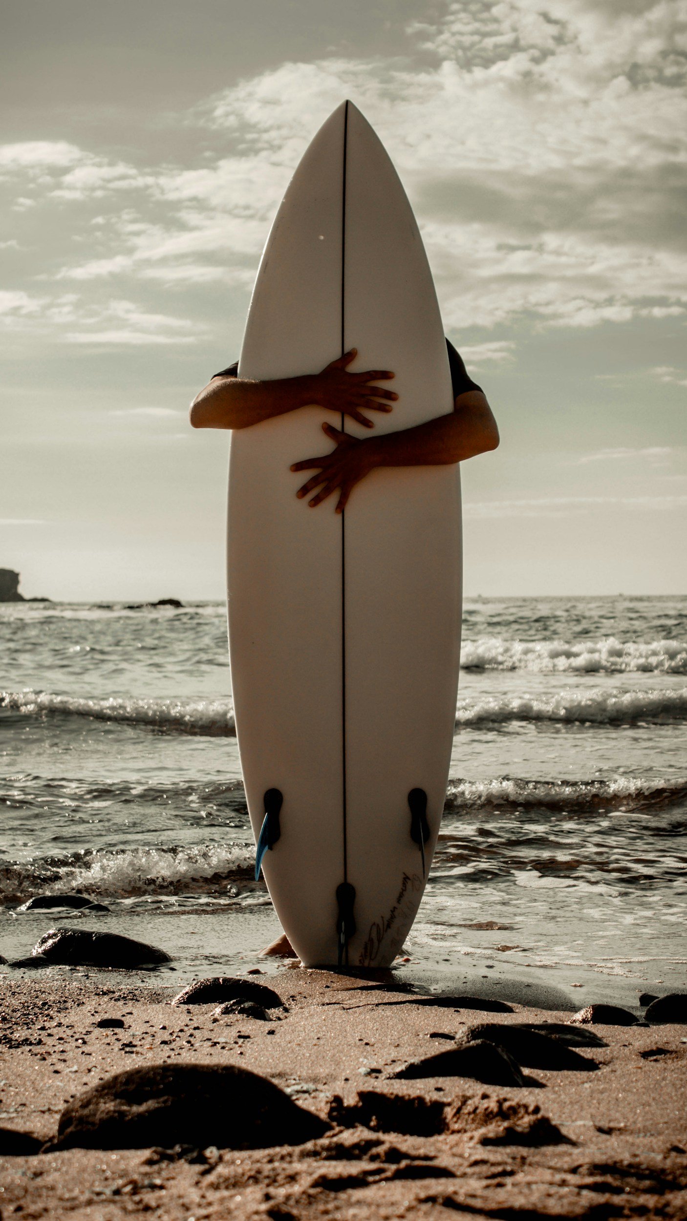 A person standing on the beach holding a surfboard, with the ocean and cloudy sky in the background.