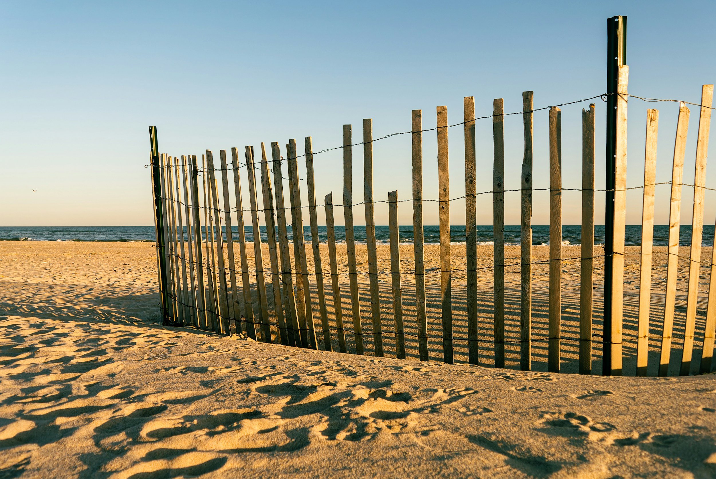 A wooden beach fence with barbed wire runs along a sandy beach with the ocean in the background. The sky is clear and blue, and the sun casts shadows on the sand.