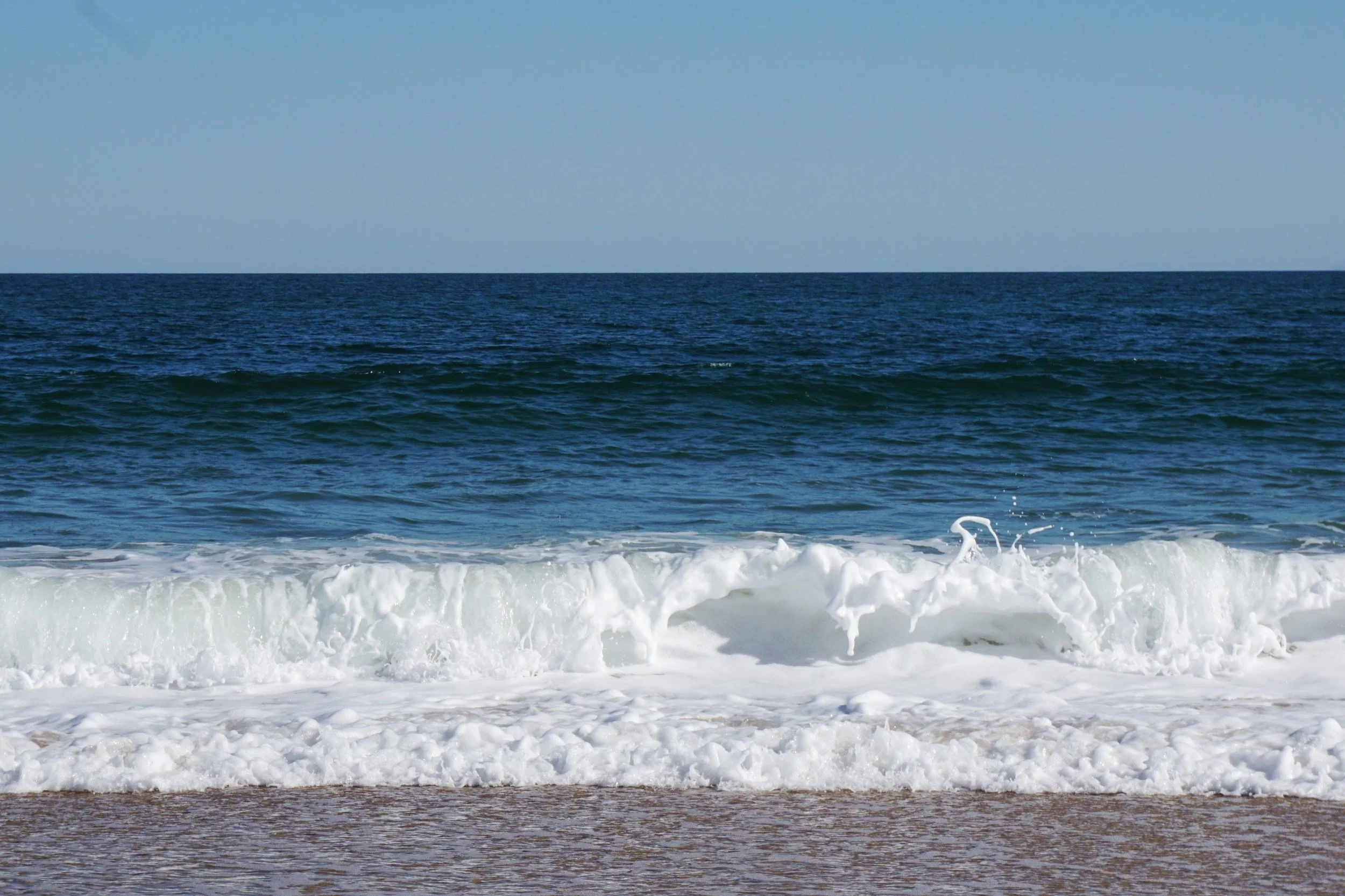 Multiple layers of the ocean with waves, transitioning from brownish near the shore to deep blue further out, under a clear sky.