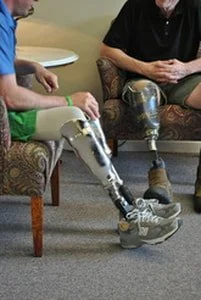 Two People with prosthetic legs sitting on a patterned armchairs, talking to each other.