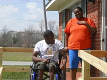 A person in a wheelchair and another person standing outside a brick building with a wooden railing, on a grassy area with trees in the background.