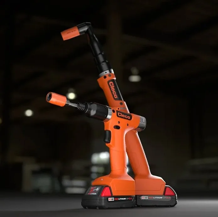 An orange cordless electric power tool, likely a screw gun or drill, with a long black barrel and an orange handle, standing upright on a dark surface with a blurred background.