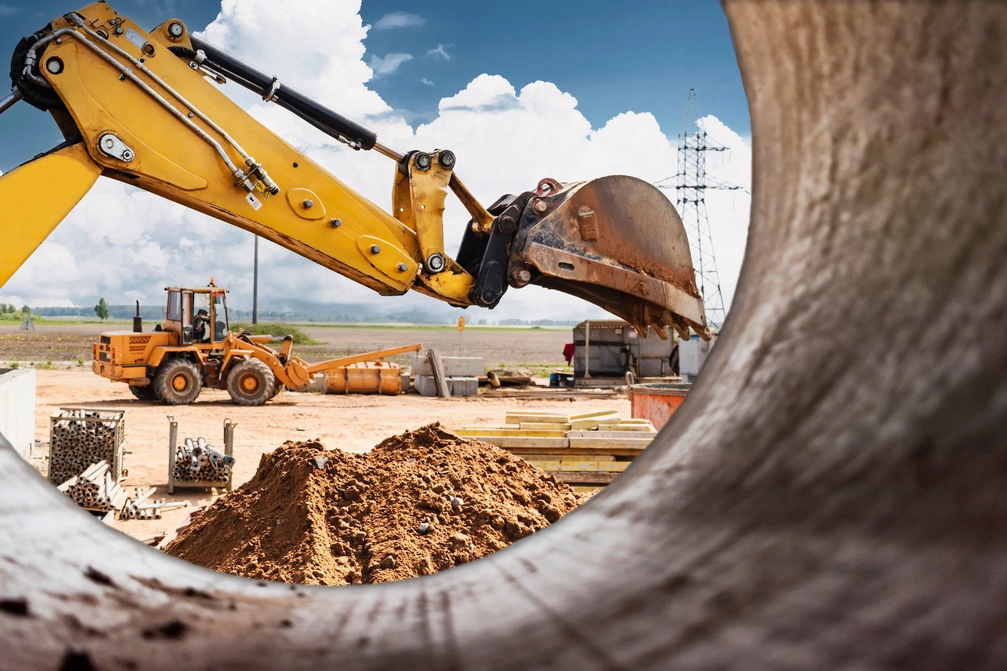 Construction site with bulldozer and excavator working on earth, pipes and construction materials scattered around, power lines in the background, under a partly cloudy sky.