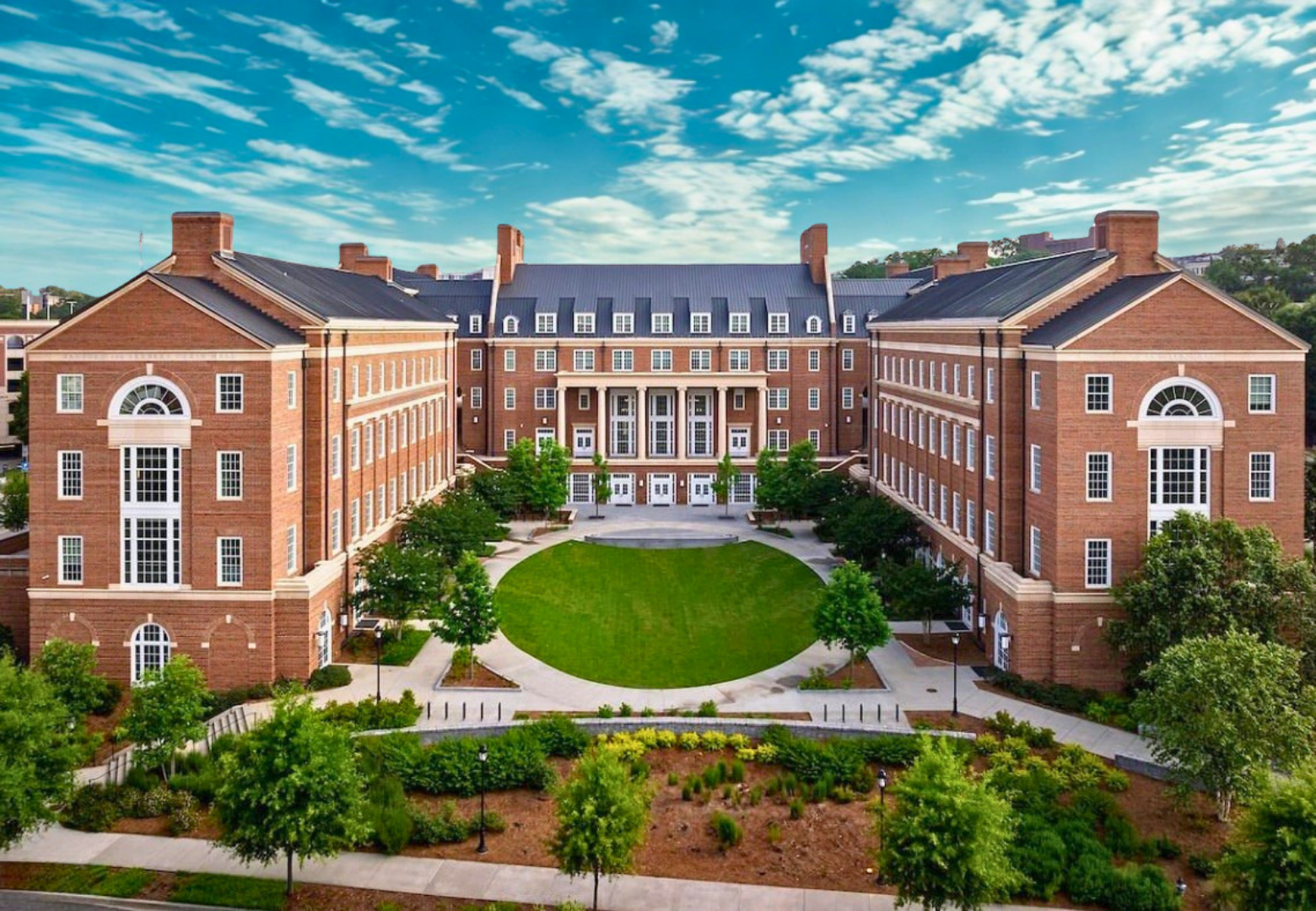 A large brick college building with multiple levels, surrounded by a green lawn and trees, under a partly cloudy sky.