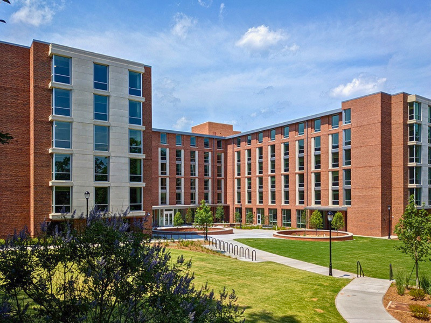 Modern brick apartment building with landscaped courtyard, green grass, trees, walking path, and lampposts under blue sky.