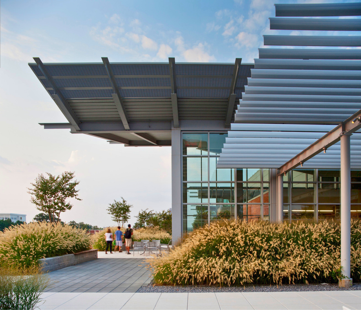 Modern building with large glass windows and metal paneling, surrounded by landscaped plants and trees, with a group of people walking outside on paved pathway under a partly cloudy sky.