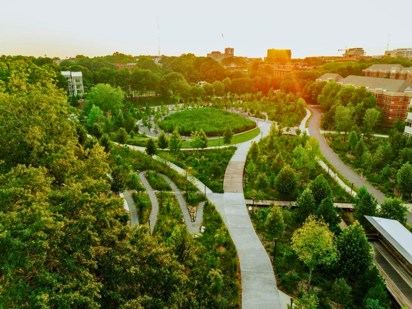 Aerial view of a park with winding pathways, green trees, and buildings in the background during sunset.