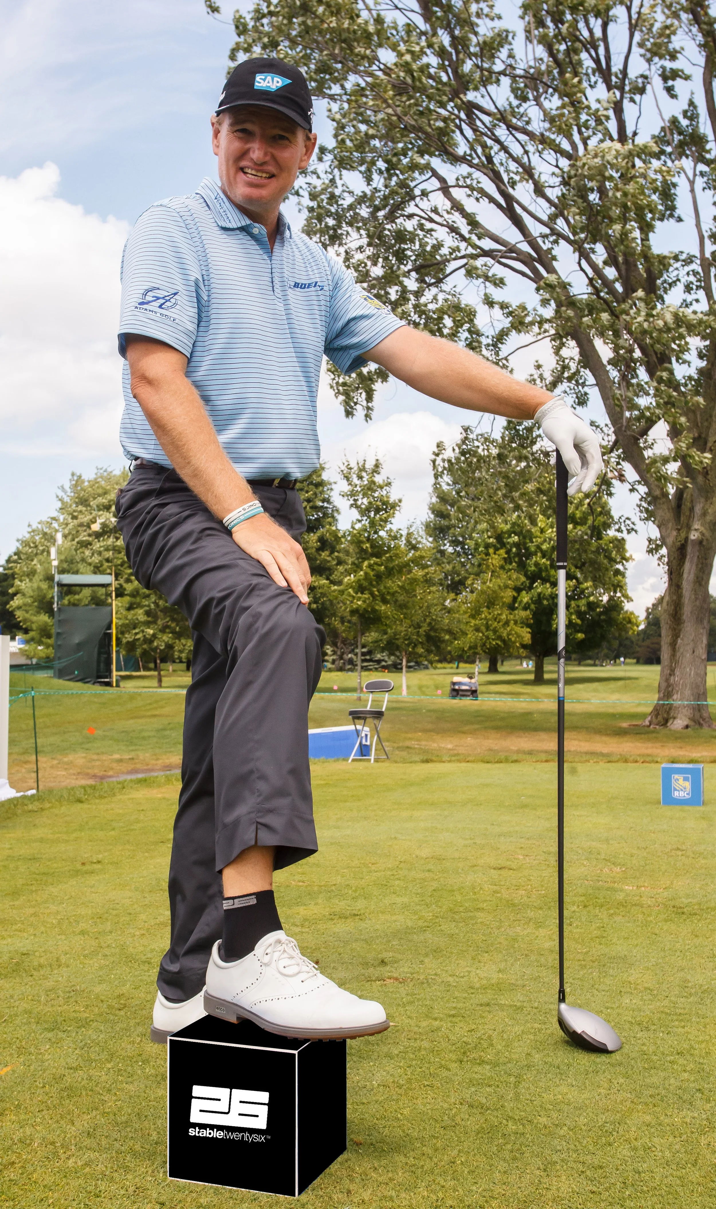 A man on a golf course, balancing on a black cube with the number 23 and the text "stablew26". He is holding a golf club in his right hand, wearing a blue cap, striped polo shirt, black pants, and white golf shoes, smiling at the camera.