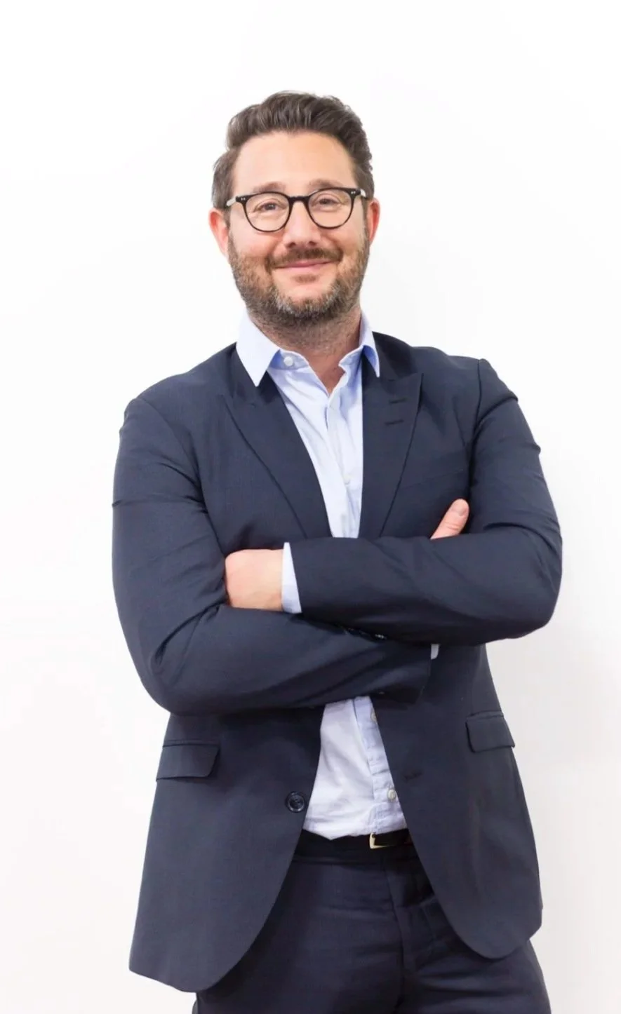 A man with glasses and a beard, wearing a dark suit and light blue shirt, standing against a white background with arms crossed, smiling.