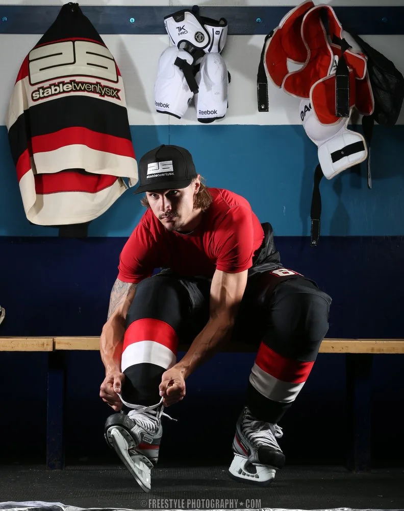 A male hockey player in a red shirt and black pants with red and white stripes, sitting on a bench, tying his ice skates. Hockey gear, including a jersey, gloves, and a helmet, hangs on hooks behind him.