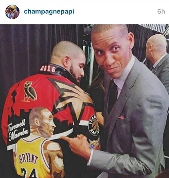 Man in a gray suit signing a Los Angeles Lakers basketball player's shoulder tattoo at an event.