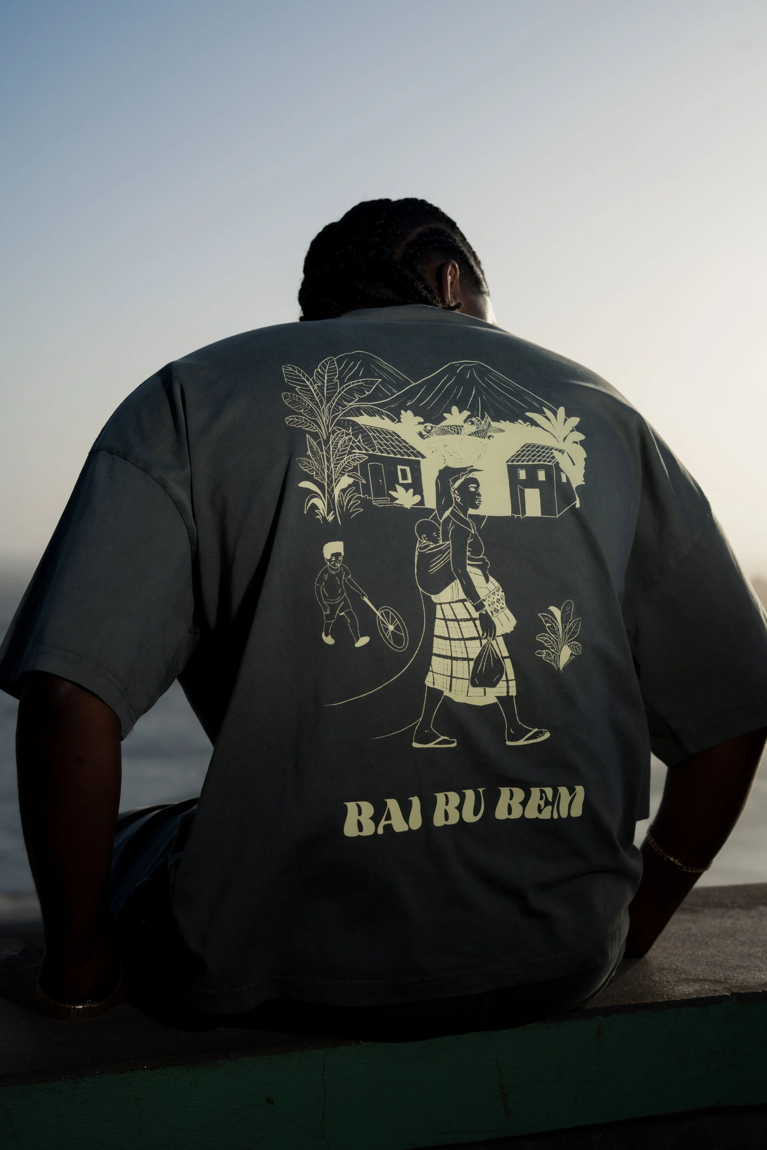 Vue de dos d'une personne assise, portant un t-shirt gris avec une illustration de village, montagnes, et personnages traditionnels, avec le texte 'Bai Bu Bem', au coucher du soleil.