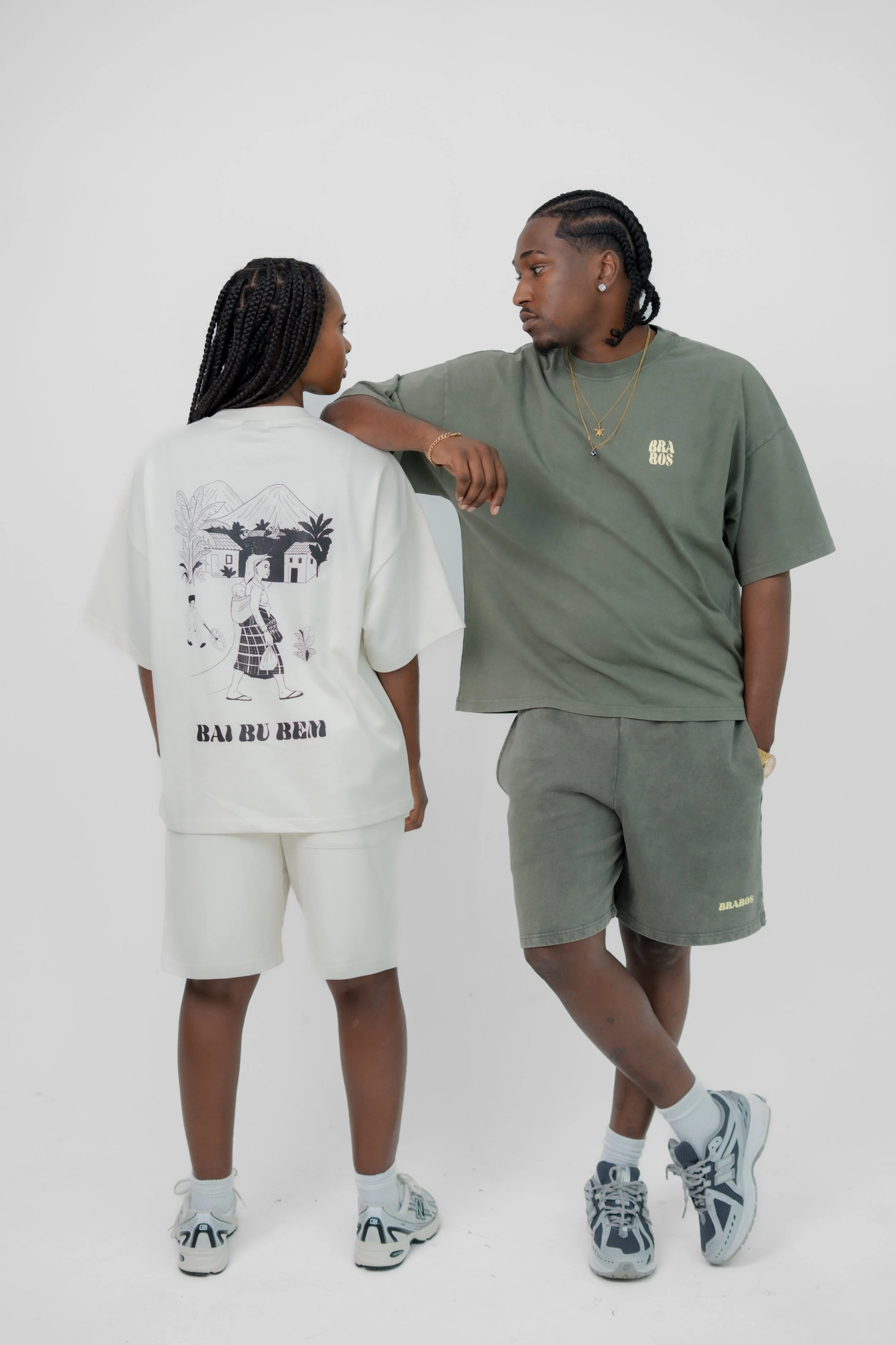Deux personnes portant des vêtements de style streetwear décontracté, avec des baskets, se tenant contre un mur blanc, la personne de gauche porte un sweat blanc avec un dessin de village et le texte 'Bai Bu Bém', tandis que la personne de droite porte un t-shirt vert avec le logo 'BRABOS'.