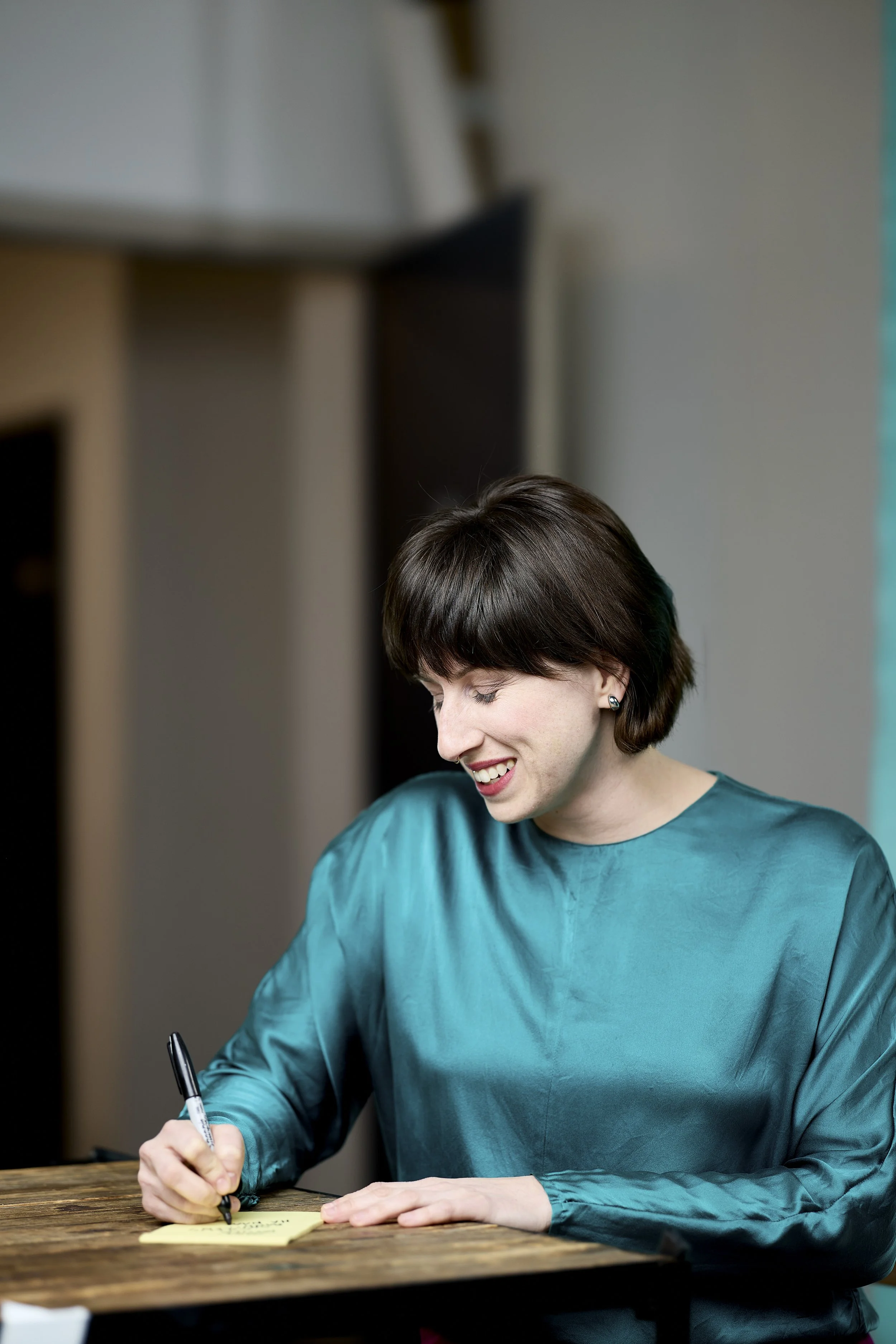A woman with short brown hair, wearing a teal long-sleeve shirt, is smiling and writing on a yellow sticky note with a black marker while sitting at a wooden table.