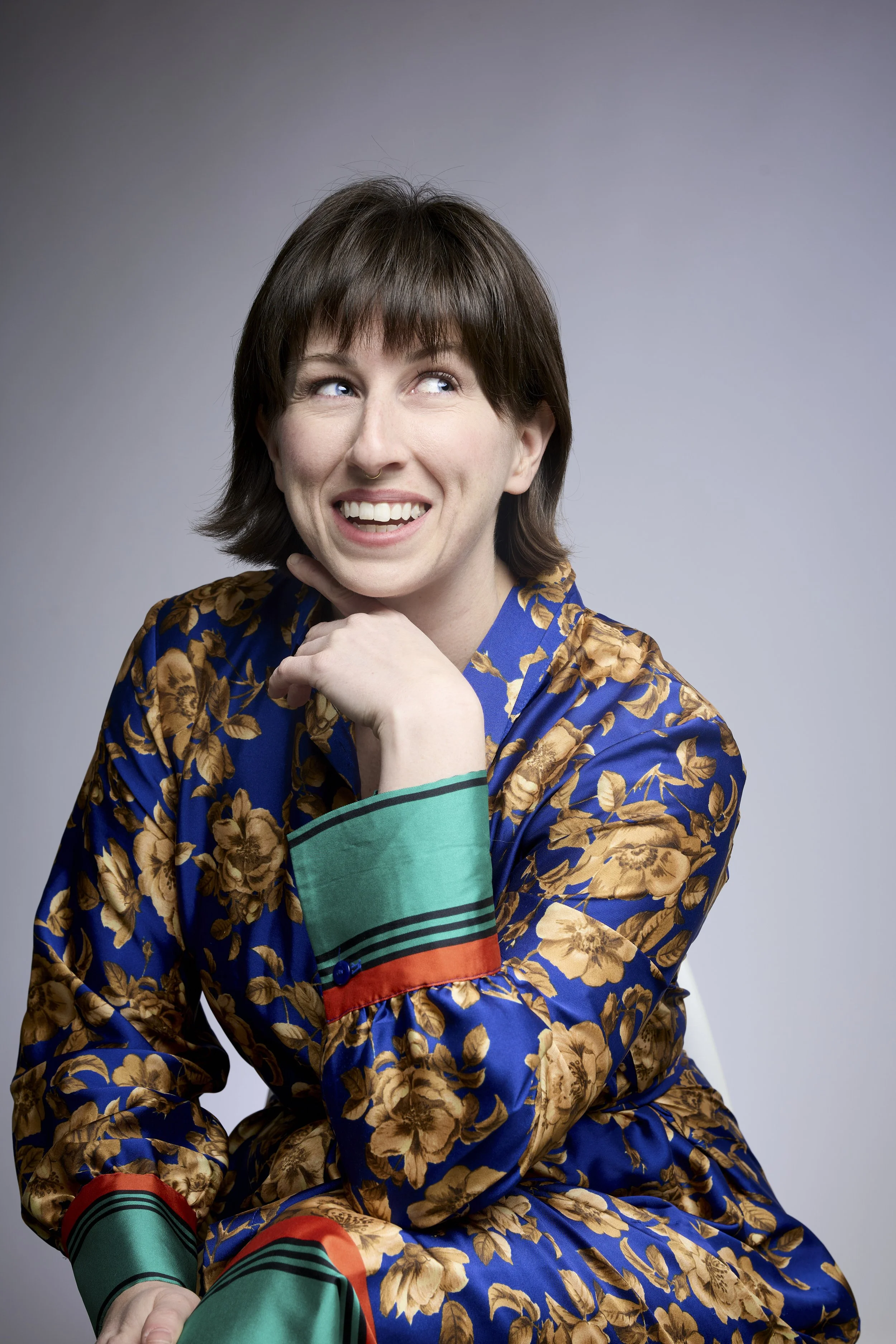 A woman with short brown hair and a wide smile, wearing a colorful floral and striped patterned blouse, looking to her right with her left hand resting on her chin, against a plain gray background.