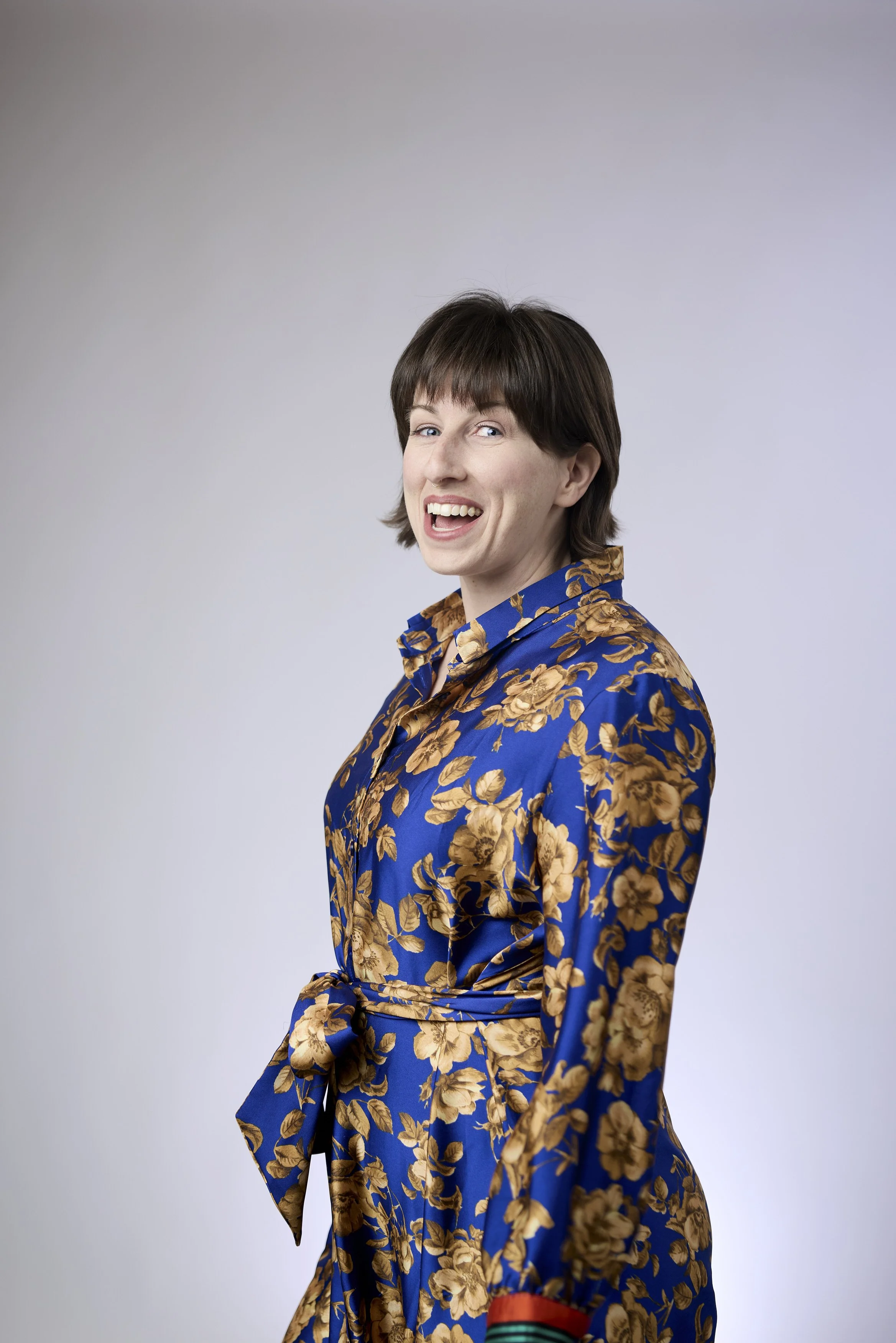 A woman with short brown hair, wearing a blue dress with a gold floral pattern and a belt tied in a knot at the waist, smiling and looking at the camera against a plain light gray background.