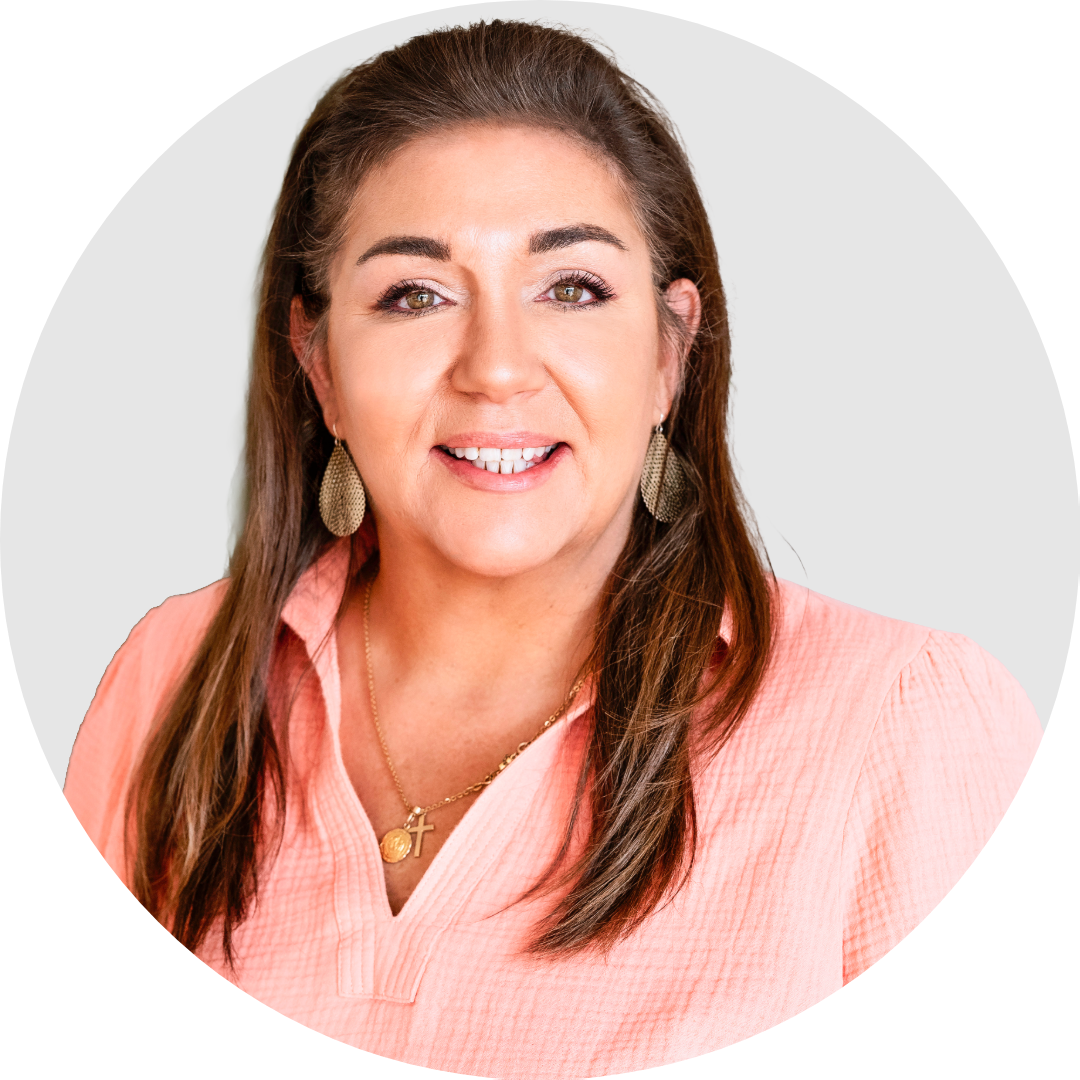 A woman with brown hair smiling, wearing a peach blouse, gold earrings, and necklaces, against a plain background.