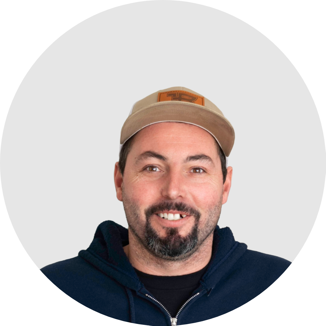 A man smiling wearing a beige baseball cap and a navy hoodie against a light gray background.