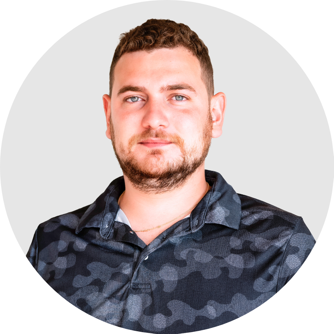 A young man with short brown hair, blue eyes, and a beard, wearing a black camouflage-patterned shirt against a plain light gray background.