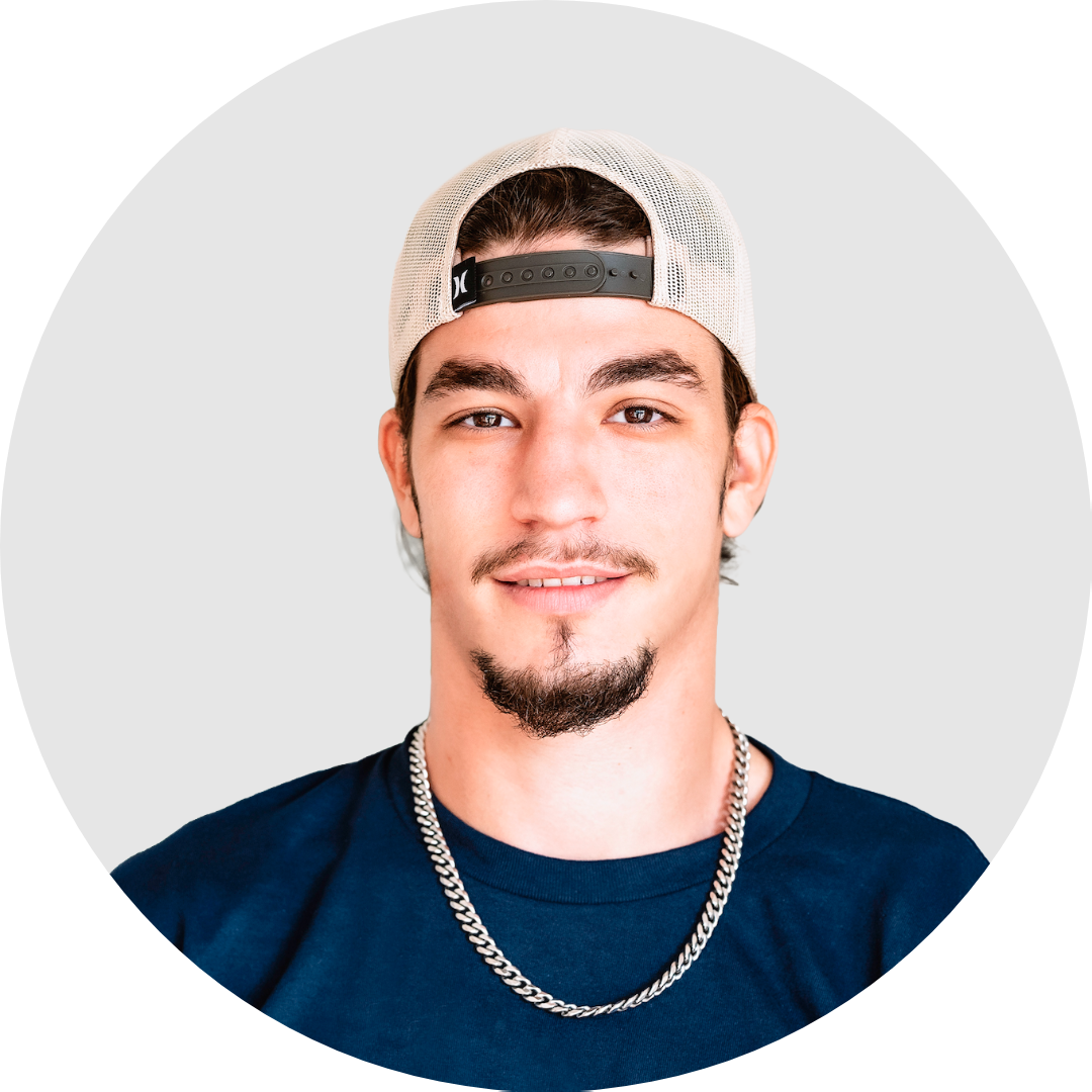 A young man with a goatee, wearing a beige baseball cap backward, a navy blue T-shirt, and a silver chain necklace, smiling at the camera.