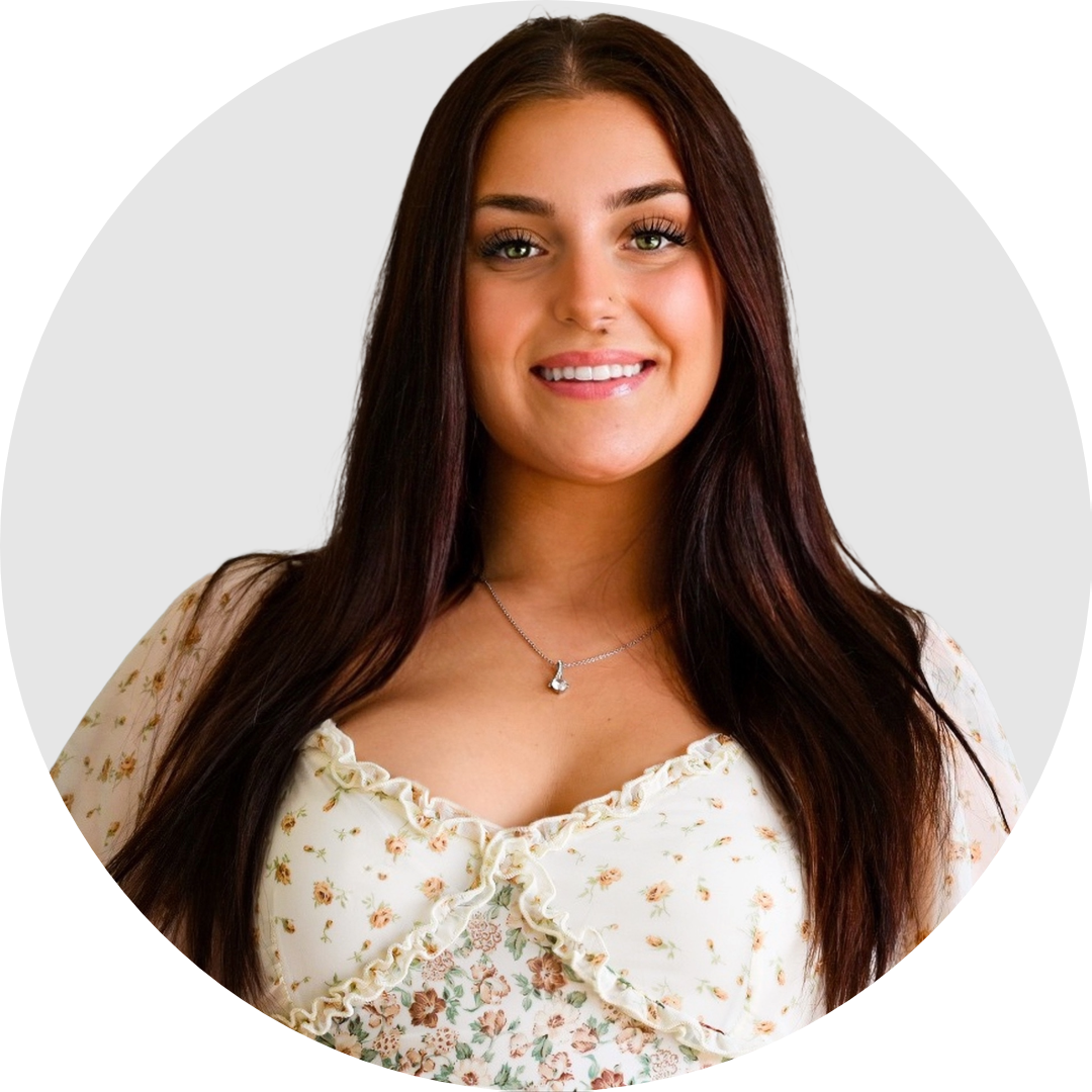 A young woman with long brown hair and light skin, smiling, wearing a floral blouse and a silver necklace, against a plain background.