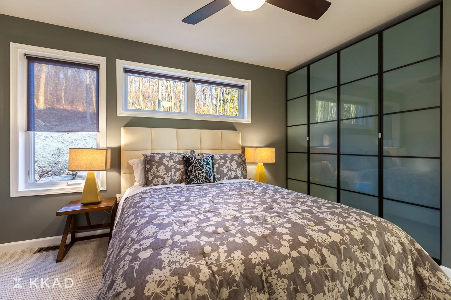 A bedroom with a bed, two lamps on wooden nightstands, a window with a view of trees, and a large wardrobe with frosted glass sliding doors.