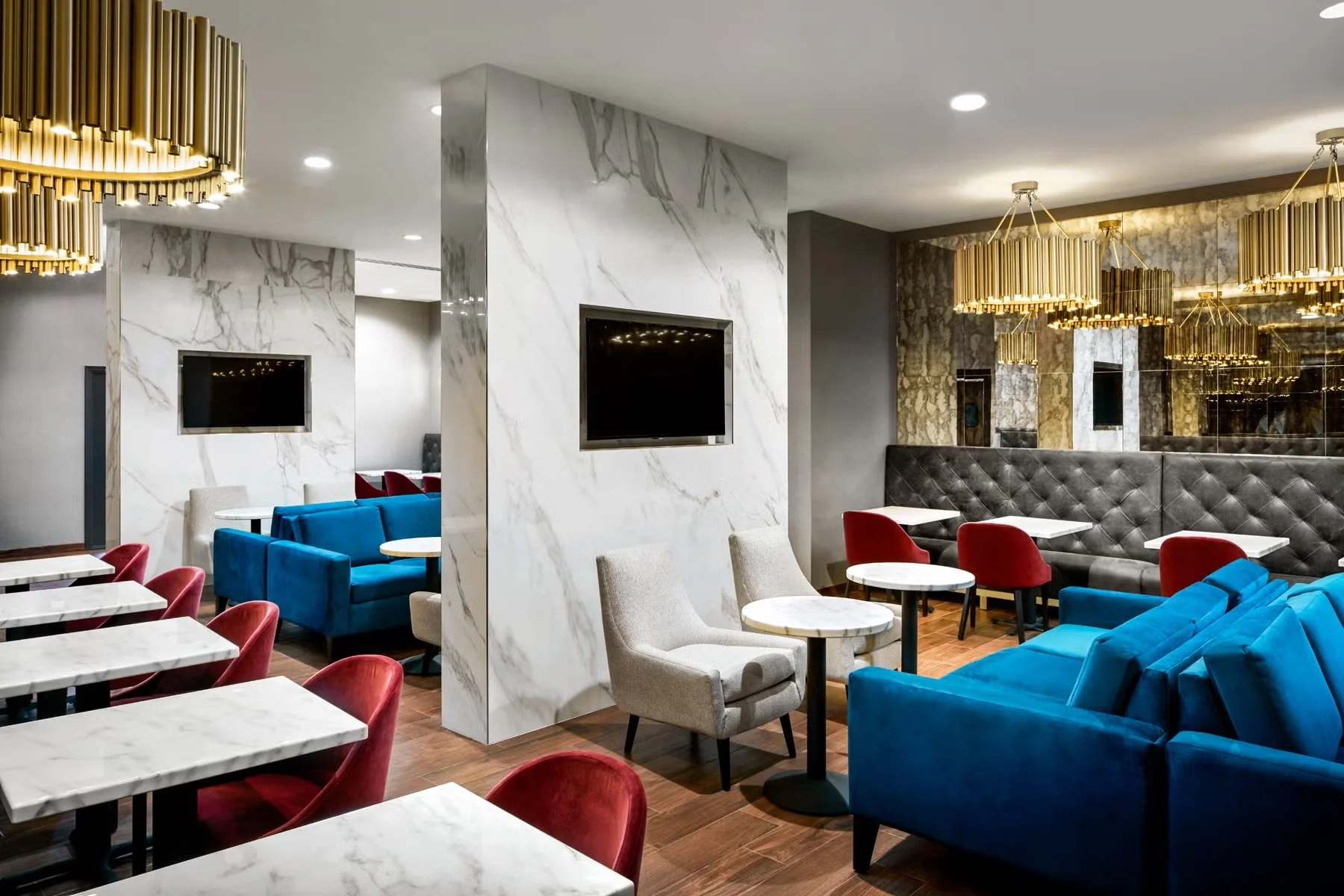 Interior of a modern lounge with colorful chairs, marble tables, white and gray walls, a gray tufted booth, and gold chandeliers.