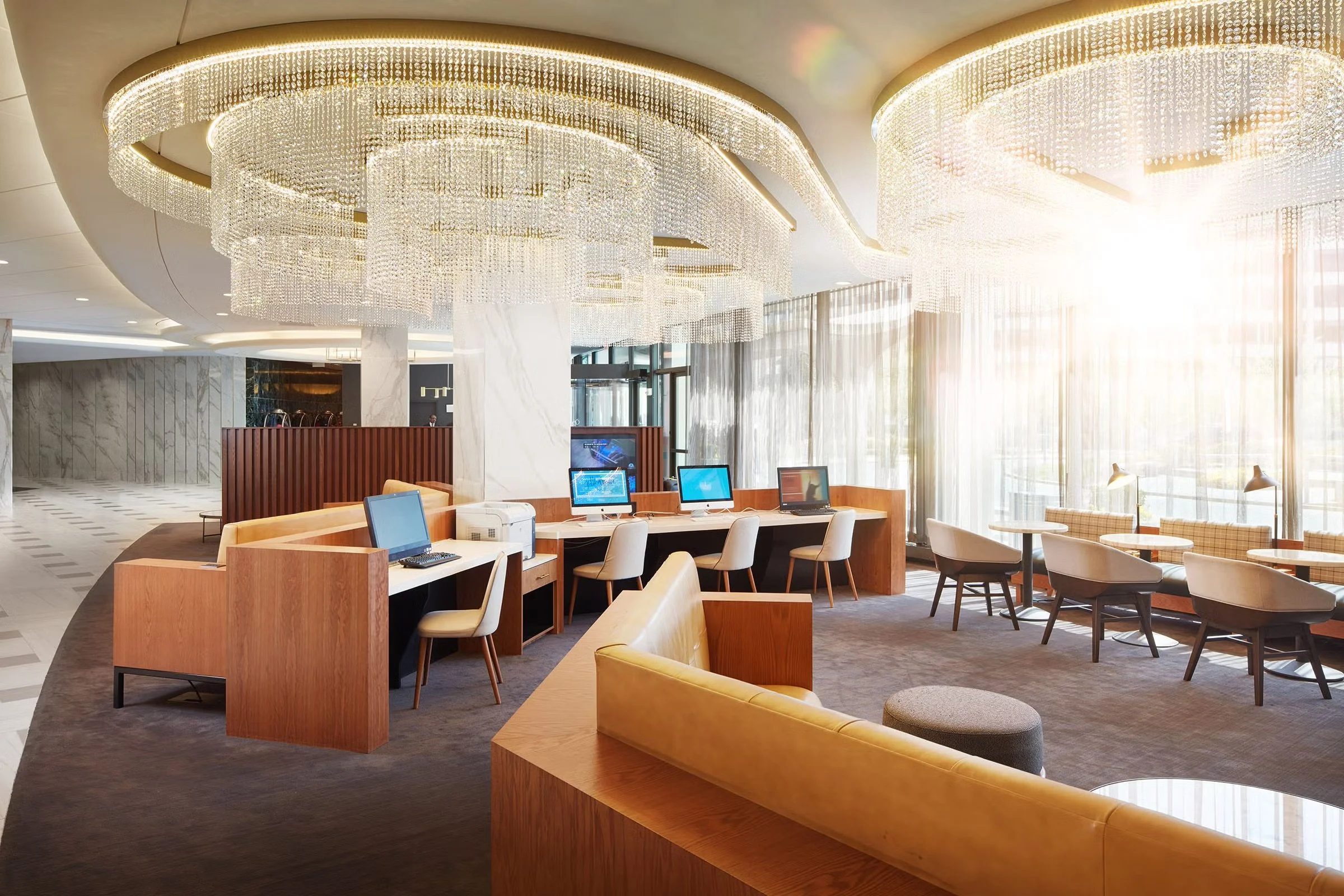Elegant hotel lobby with a large, ornate crystal chandelier, check-in desk with computers, and seating area with tables and chairs near large windows with curtains, bright sunlight streaming in.