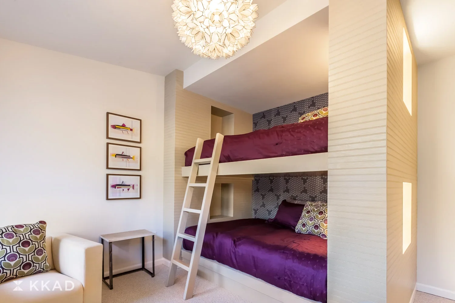 A bedroom featuring a white bunk bed with purple bedding and patterned pillows. The room has a decorative wall with dark star-patterned wallpaper behind the beds, three colorful framed pictures on a white wall, a cream-colored armchair with a pattern
