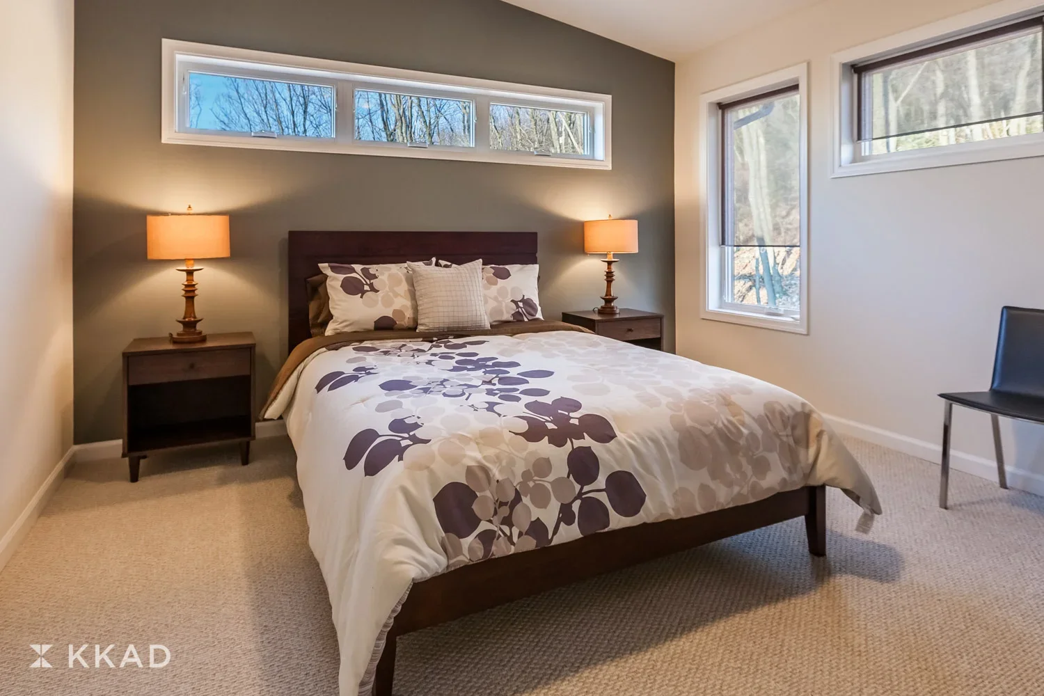 A bedroom with a bed, two nightstands with lamps, and three windows showing trees outside.