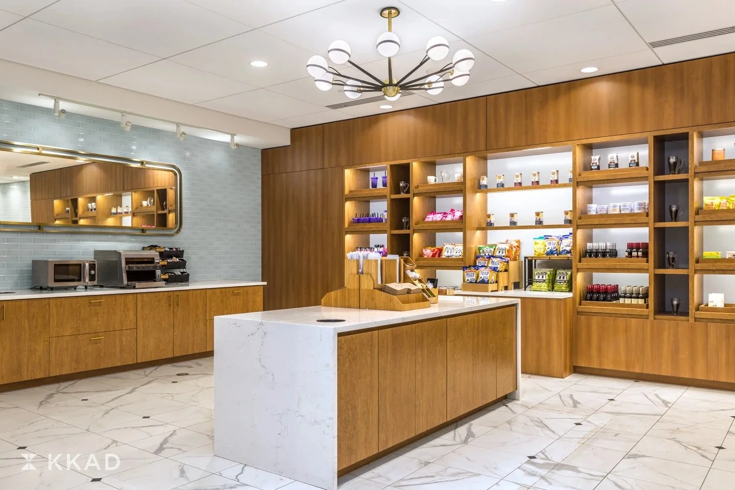 Interior of a modern coffee shop or cafe with wooden shelves stocked with snacks and condiments, a white marble counter, a microwave, and a coffee station, illuminated by contemporary lighting fixtures.