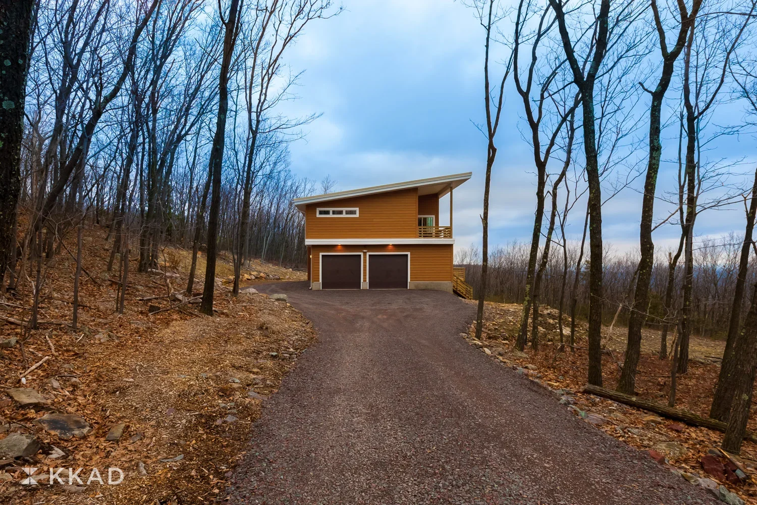 A modern two-story house with a sloped roof, situated on a wooded hill with a gravel driveway in the foreground, during early evening with overcast sky.