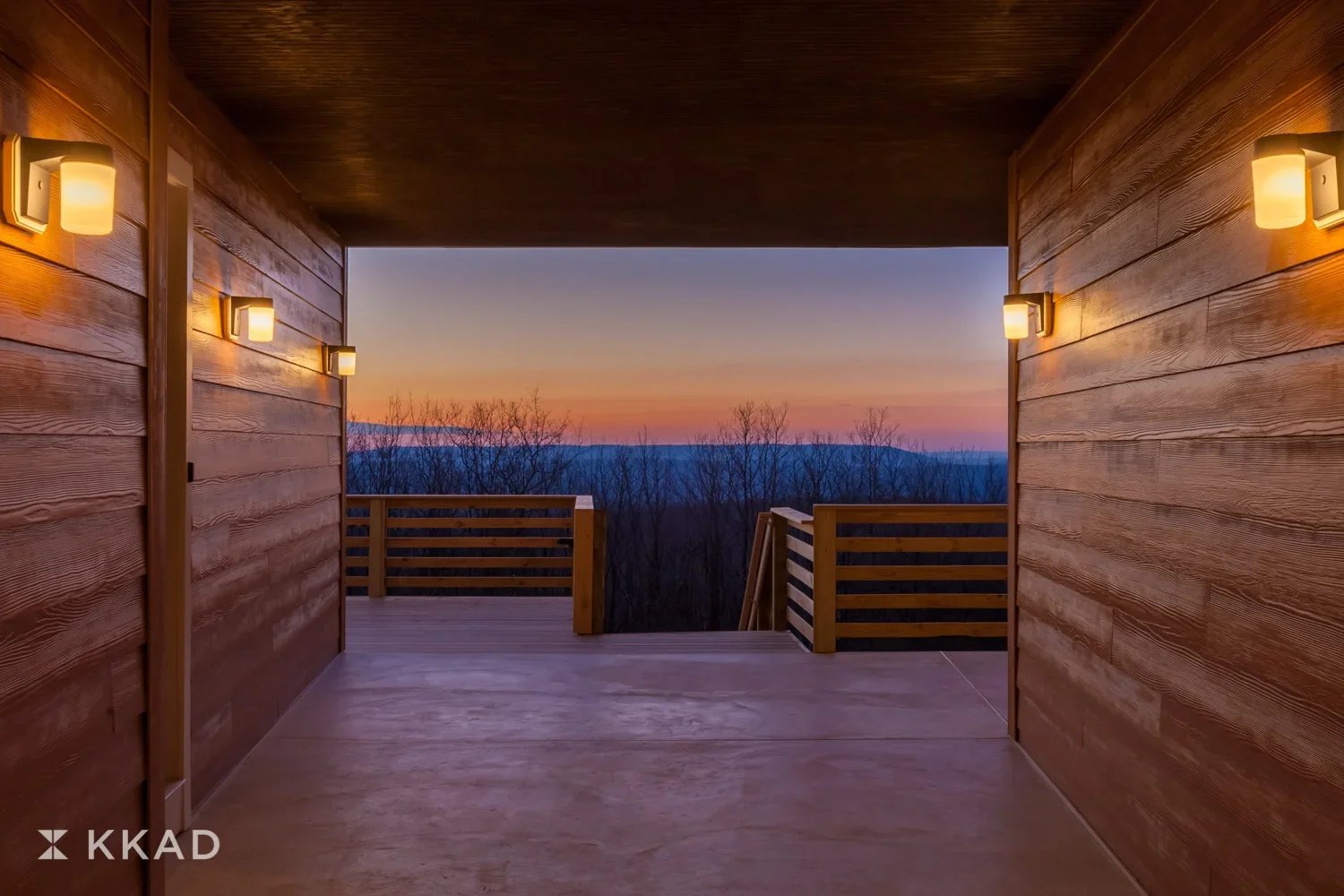View from a wooden balcony overlooking a sunset with a distant mountain range and leafless trees.