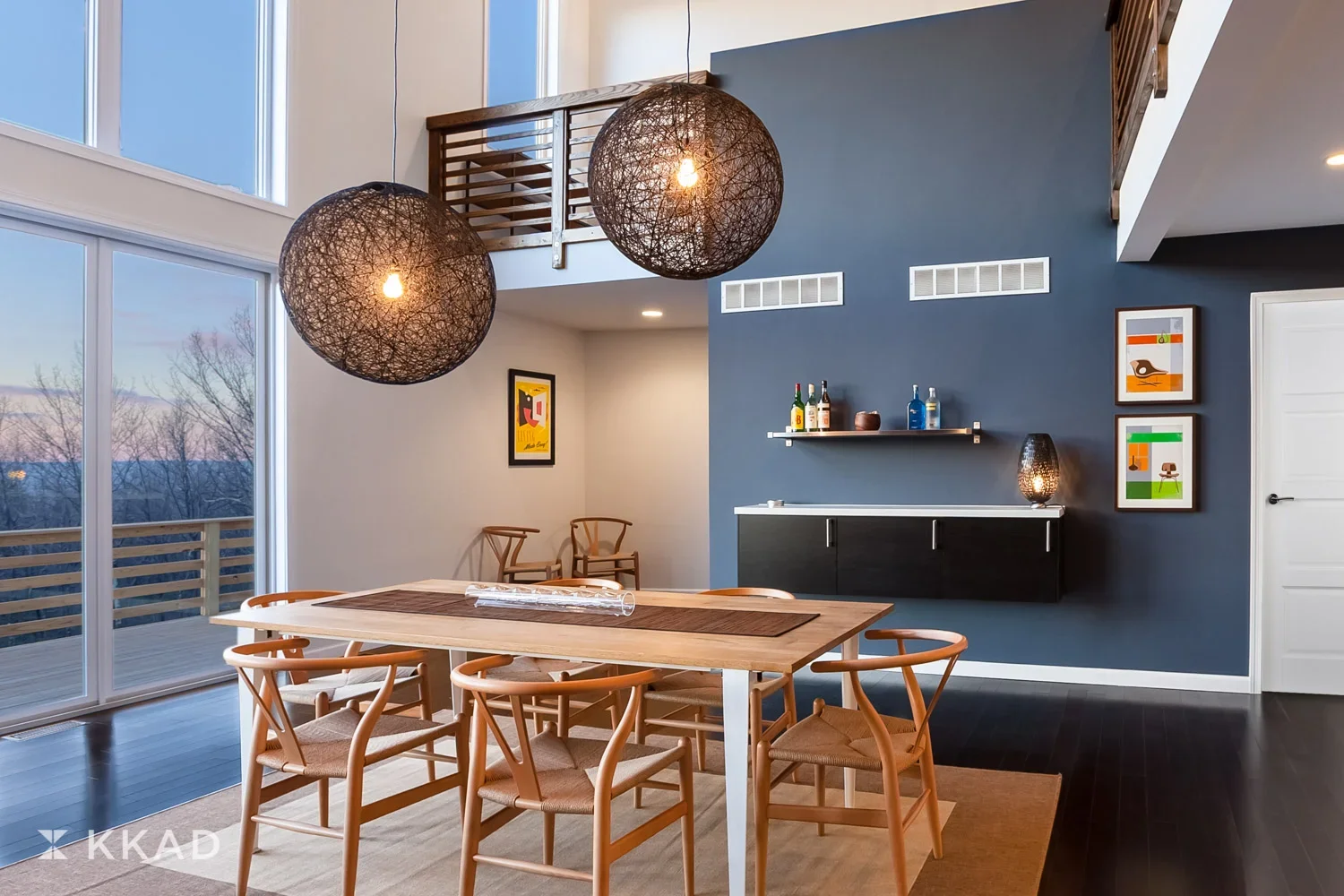 Modern dining room with a light wooden table, six matching chairs, large sliding glass door with a balcony outside, dark floor, and a navy blue accent wall with framed artwork and a floating shelf with bottles. Two large woven pendant lights hang abo