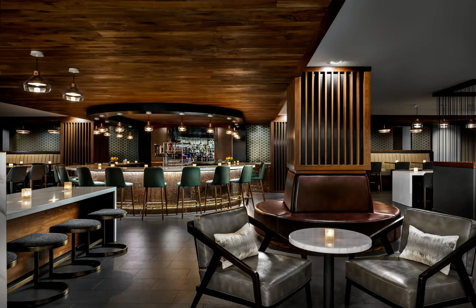 Interior of a modern bar and lounge area with a wooden ceiling, a curved bar counter with green bar stools, and various seating options including chairs and booths, illuminated by warm lighting.
