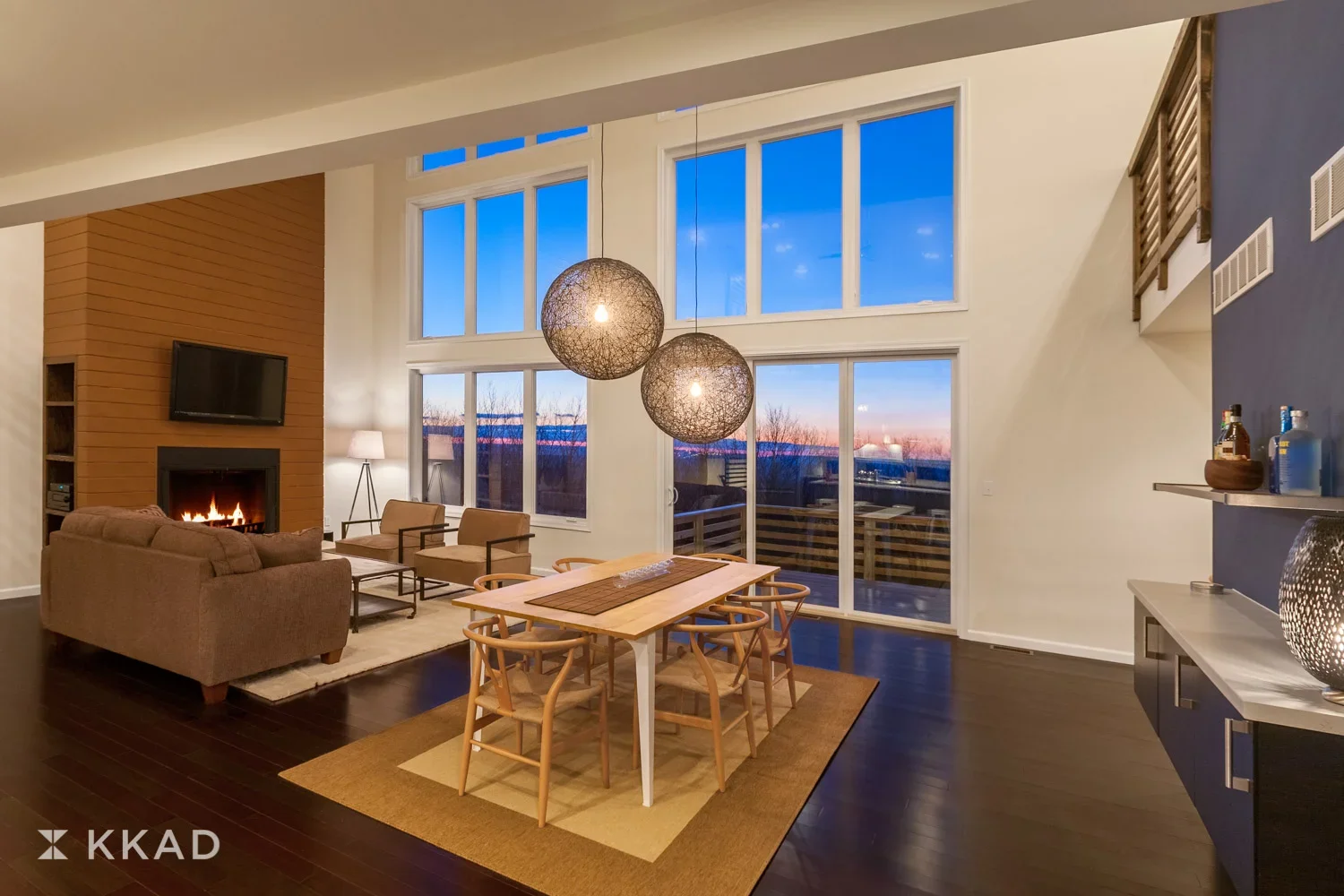Modern living room with large windows showing a sunset, a fireplace on the left, a beige sofa with chairs around it, a wooden dining table with six chairs, a dark wood floor, and a blue accent wall on the right with a shelf and decorative items.